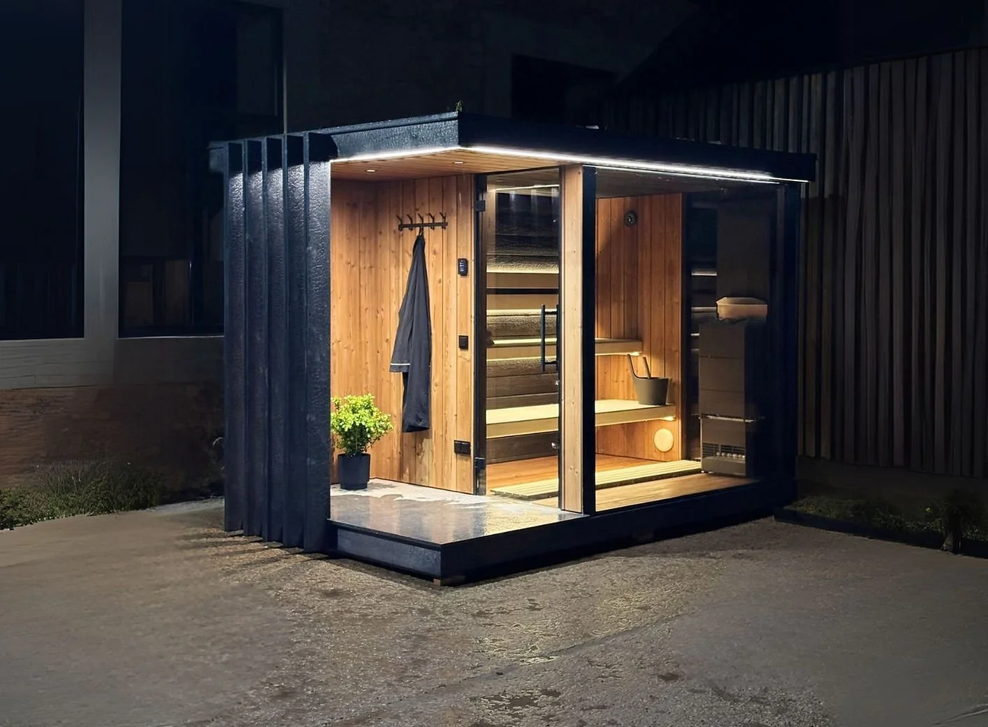 Night view of a small, modern sauna with wooden interior and exterior, illuminated at night, with a small step leading up to the entrance, a plant beside the door, and a black coat hanging on a wall hook outside.