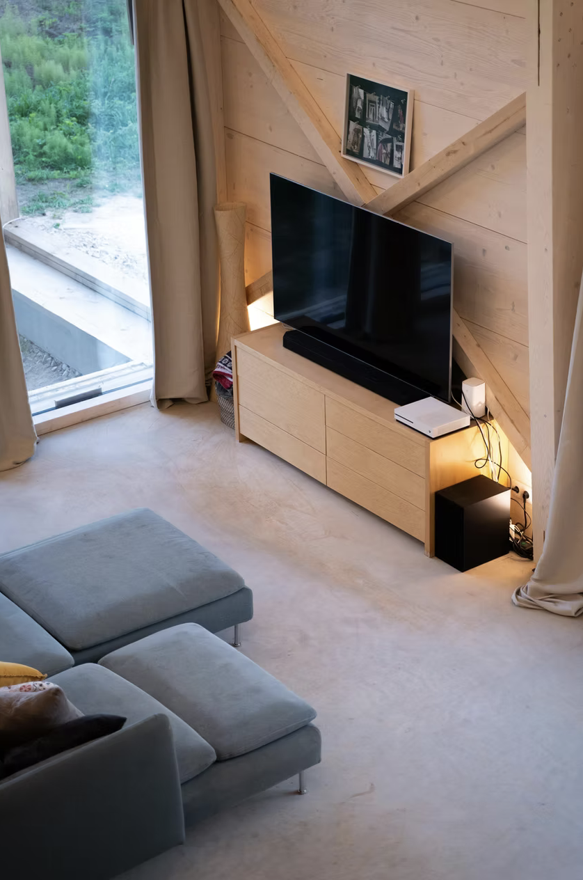 Living room with a large flat-screen TV on a light wood cabinet, a small black subwoofer, and a white gaming console, next to a window with beige curtains and outdoor greenery visible.