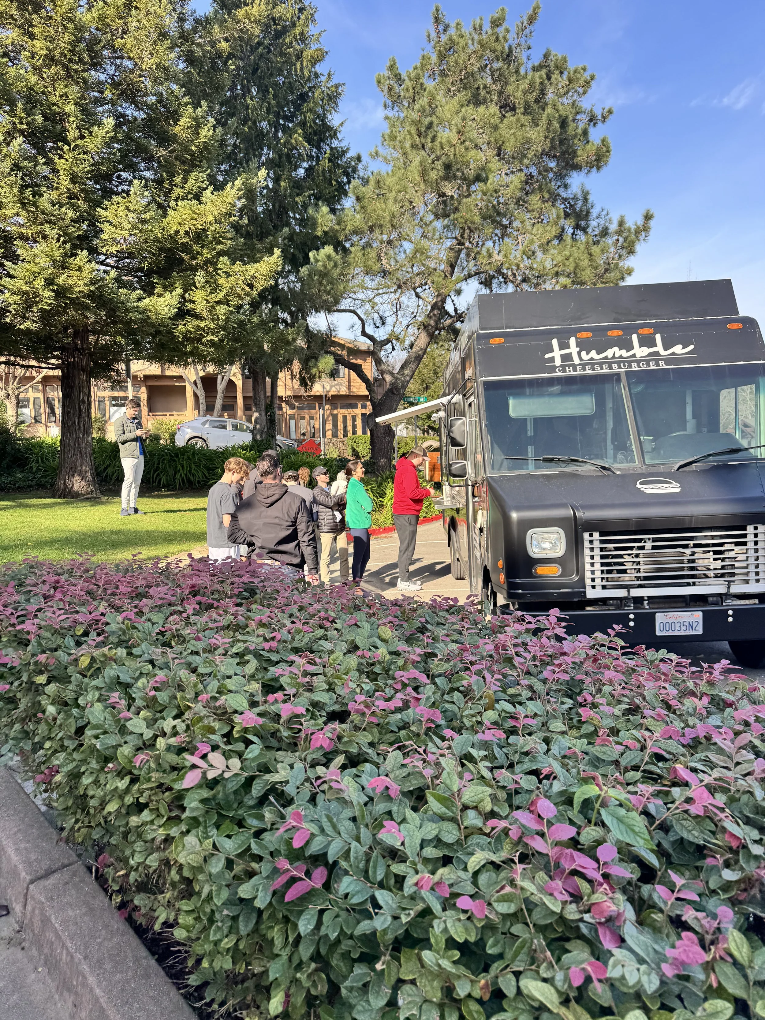 A line of people stands outside a black food truck labeled 'Humble Cheeseburger' under trees with green foliage and a blue sky.