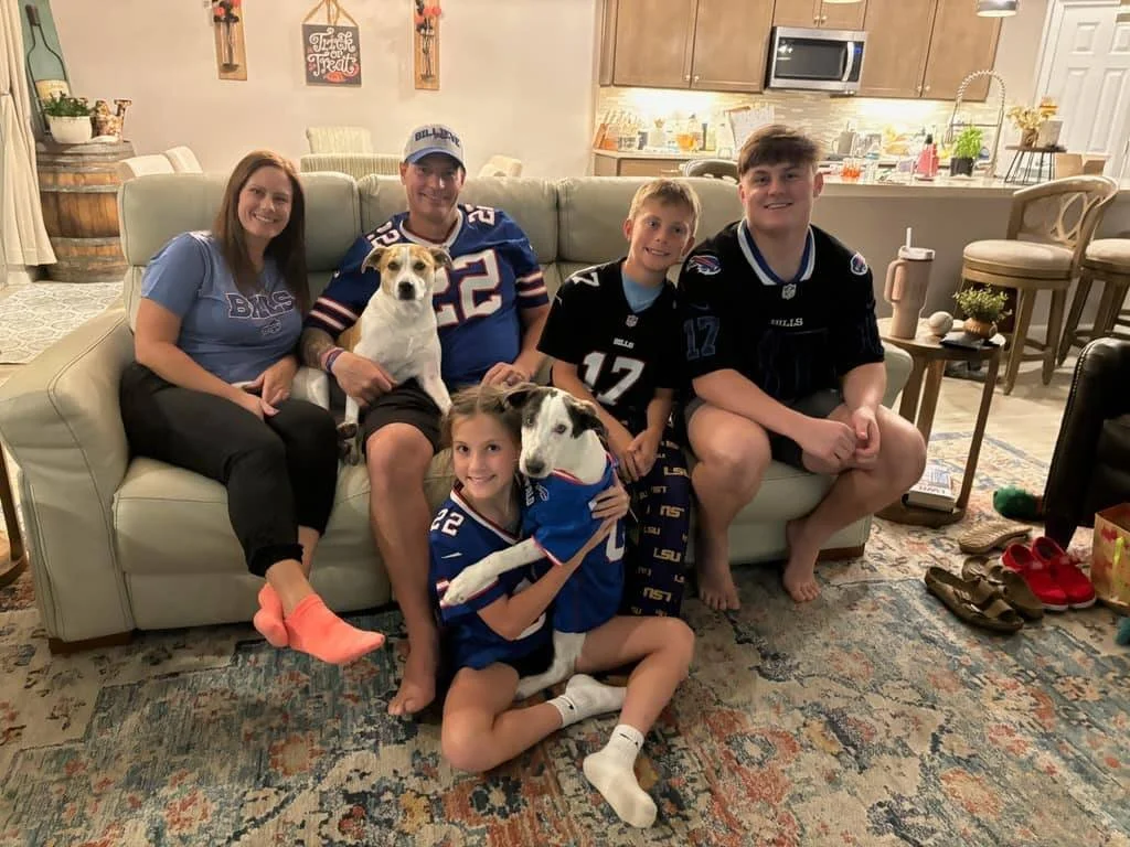 A family of five and two dogs sitting together on a sofa in a cozy living room. The family includes three children, a woman, and a man. The children and woman are all wearing sports jerseys, and the dogs are also dressed in football jerseys. The background shows a kitchen area with various decorations and furniture, and the floor has a patterned rug.