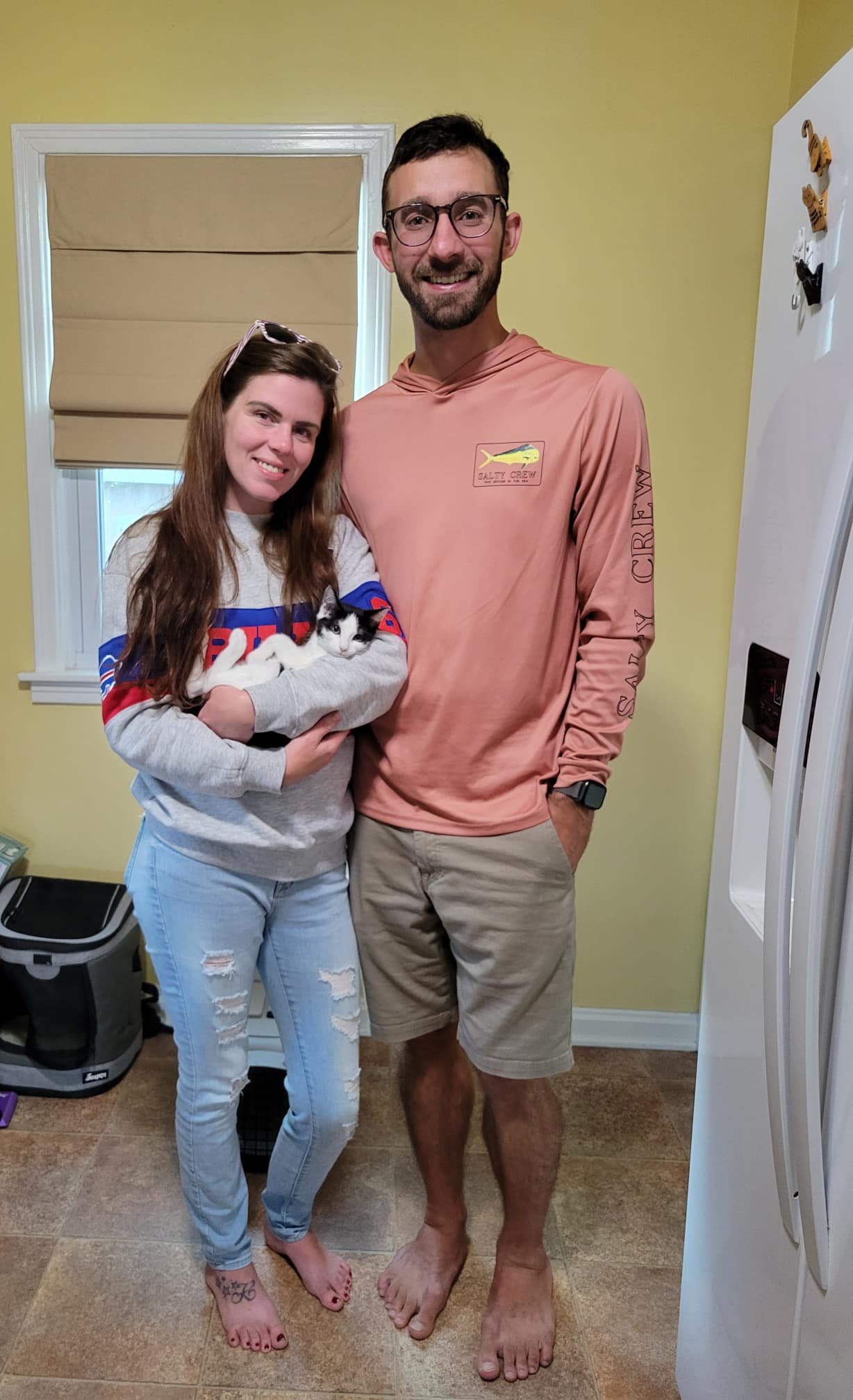A woman with long brown hair holding a black and white cat, standing next to a man with glasses, short dark hair, and a beard, both smiling indoors in front of a yellow wall and window, with a white refrigerator to the right.