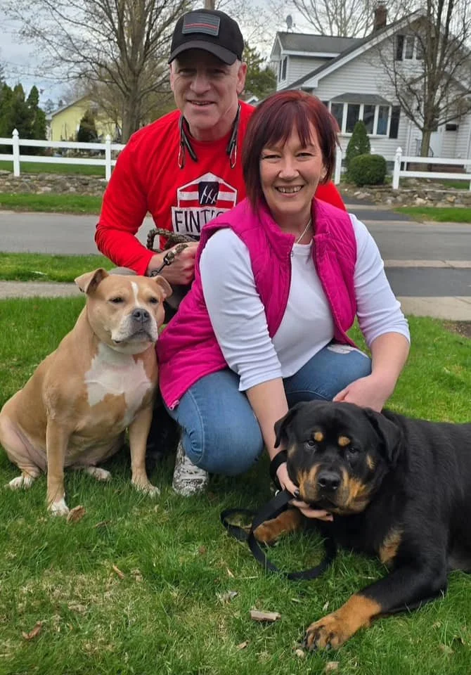 A smiling woman kneels on the grass with two dogs, one sitting and one lying down, while a man stands behind her with one hand on her shoulder, in a suburban neighborhood with houses and trees in the background.