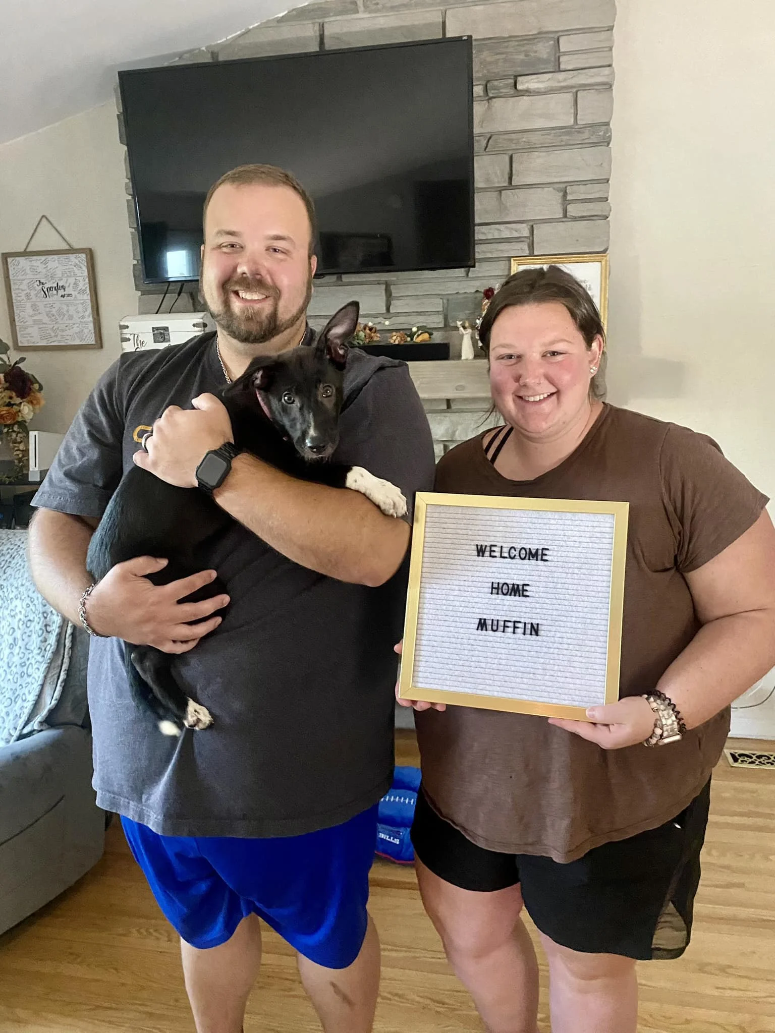 A smiling man holding a black and white puppy and a smiling woman holding a sign that reads "Welcome Home Muffin," inside a cozy living room with a stone fireplace and a large television.