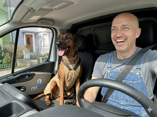 Ein Mann fährt im Lkw, neben ihm sitzt ein Hund auf dem Beifahrersitz, beide lachen.