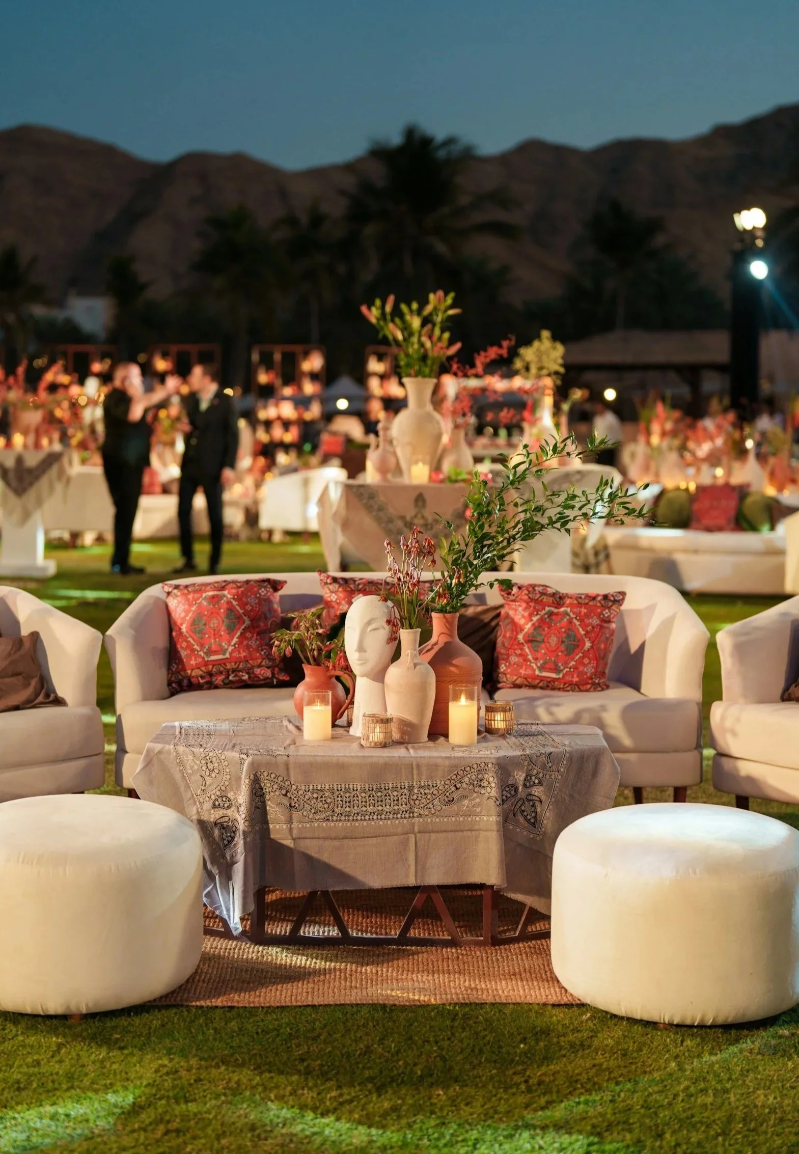 Elegant outdoor event setup with white couches, patterned pillows, a table with vases and candles, and string lights in the background at night.