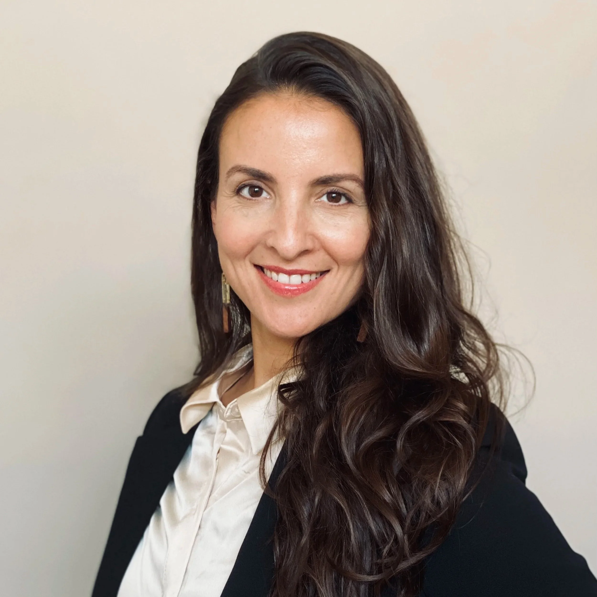 A woman with long, wavy brown hair and brown eyes, smiling, wearing earrings, a white blouse, and a black blazer, standing in front of a plain light-colored wall.