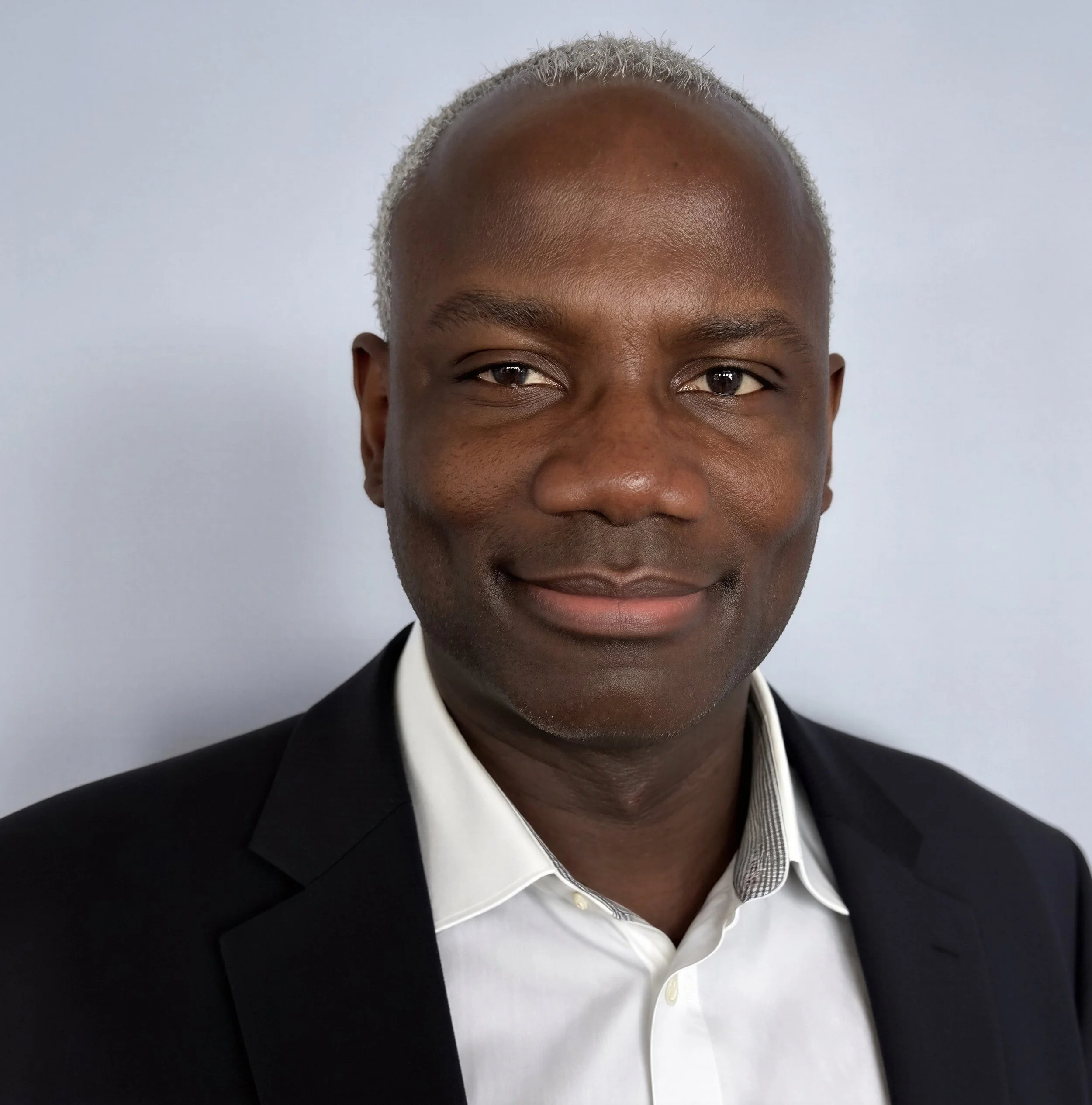 A man with dark skin and short, grey hair smiling, wearing a black suit jacket over a white collared shirt.