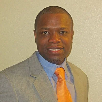 A man wearing a light gray suit, light blue shirt, and orange tie, standing against a plain light-colored wall.