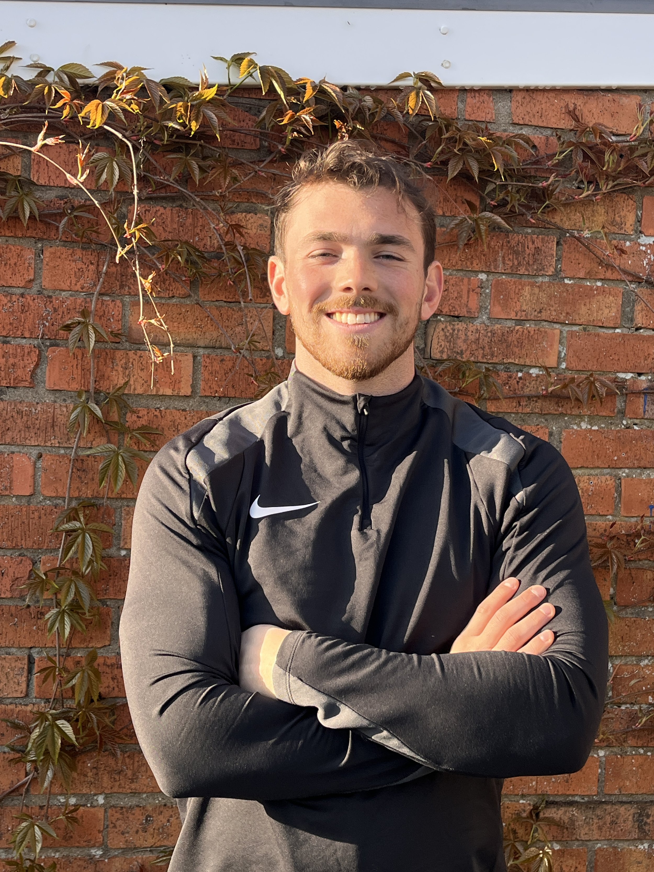 Young man smiling with crossed arms in front of a brick wall with climbing plants.