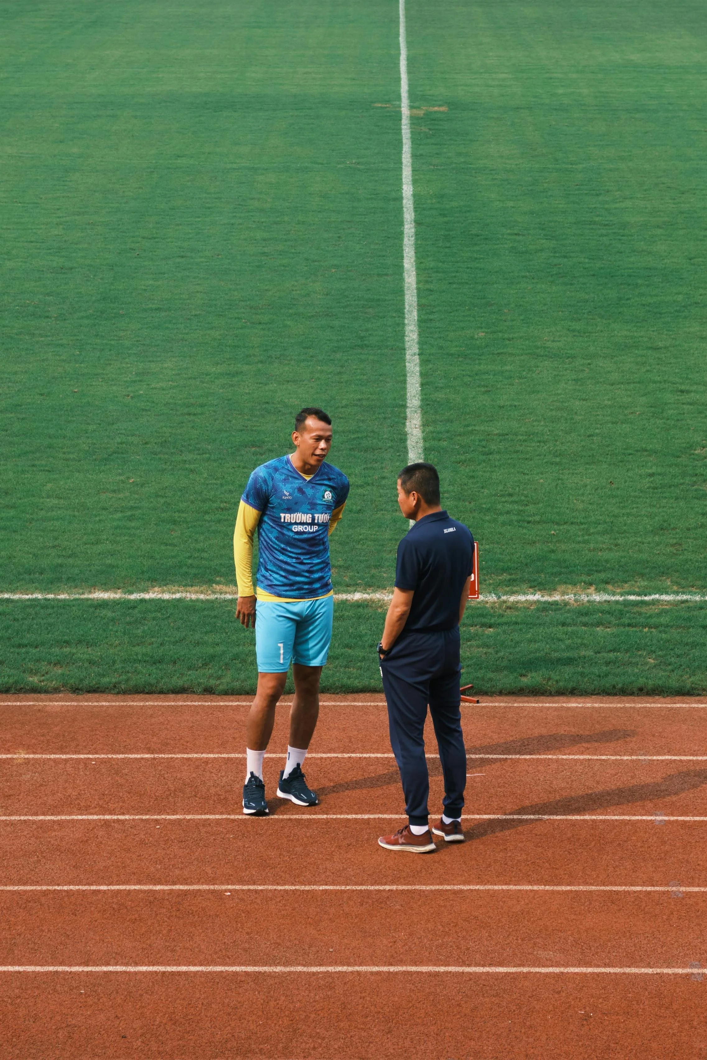 Two men standing and talking on a sports track near the grass playing field, one wearing a blue sports jersey and shorts, and the other in dark clothing.