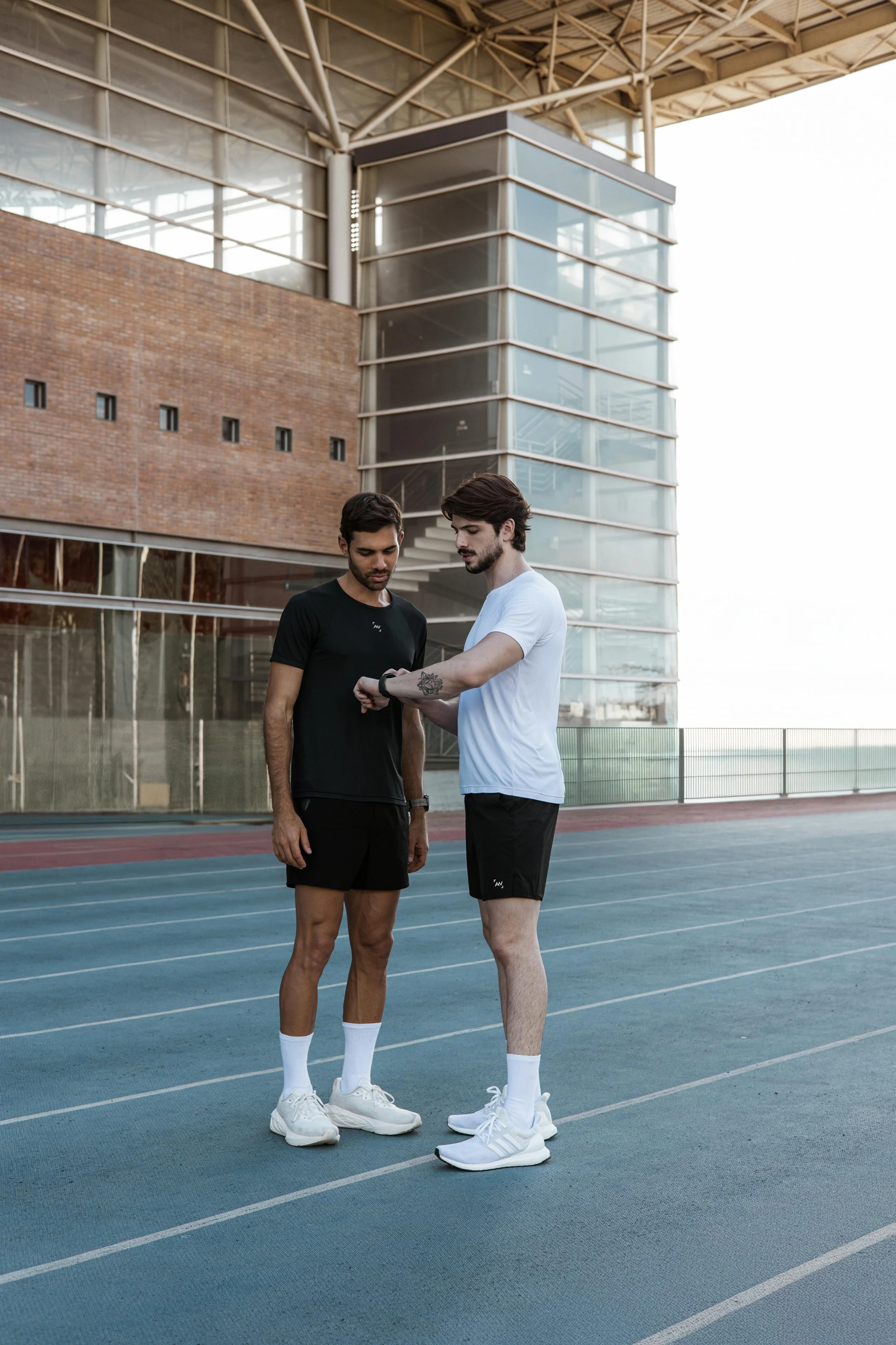 Two young men in athletic clothing standing on a running track outside a modern building, looking at a smartwatch.