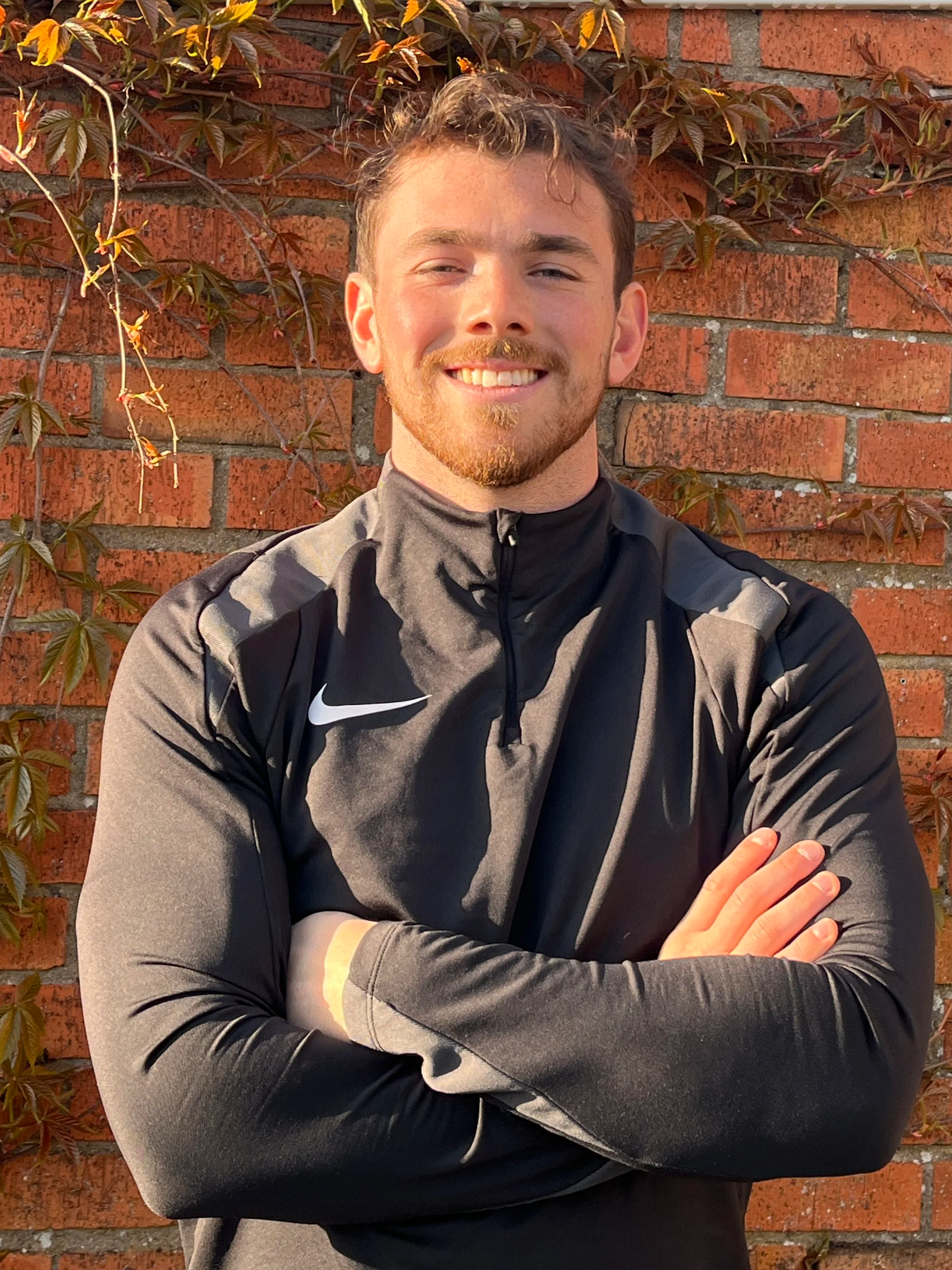 A young man with short curly hair and a beard, smiling with arms crossed, standing front of a brick wall with climbing vines.