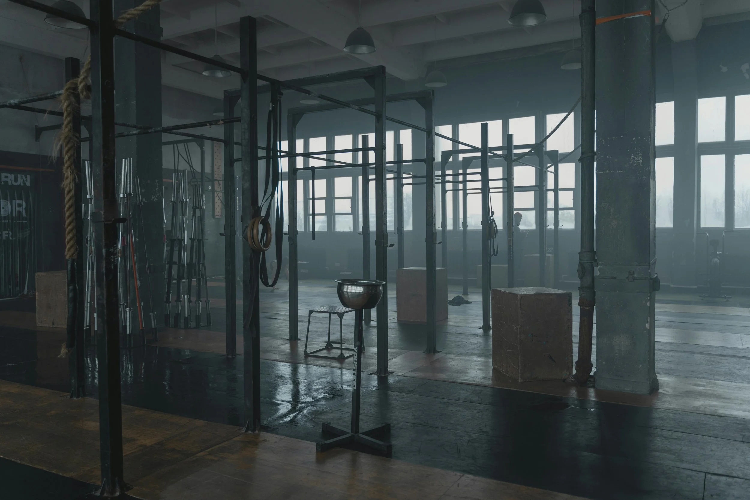 Empty gym with workout equipment, including pull-up bars, ropes, and plyometric boxes, in a spacious, industrial-style room with large windows.