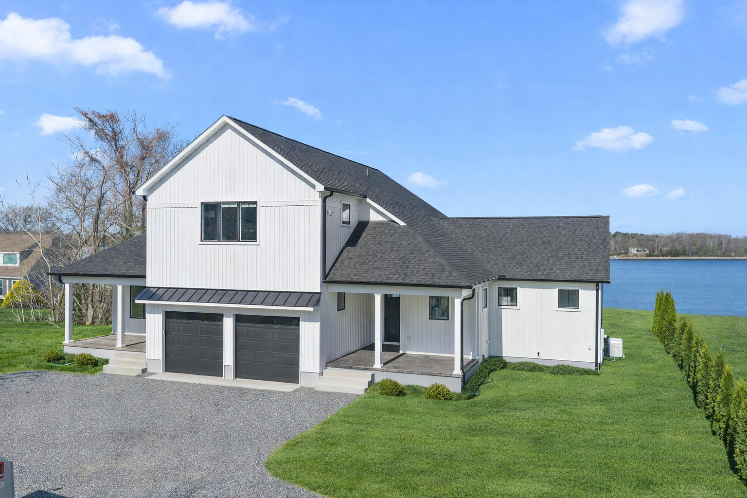 Modern white two-story house with a black roof, a two-car garage, and water in the background.