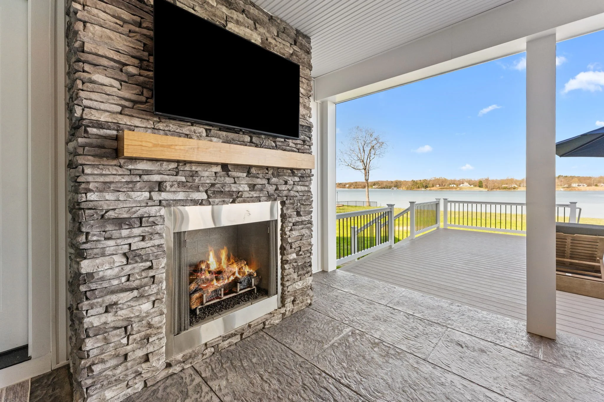 Outdoor porch with stone fireplace, mounted TV, and retractable screens opening to an expansive deck overlooking the water.