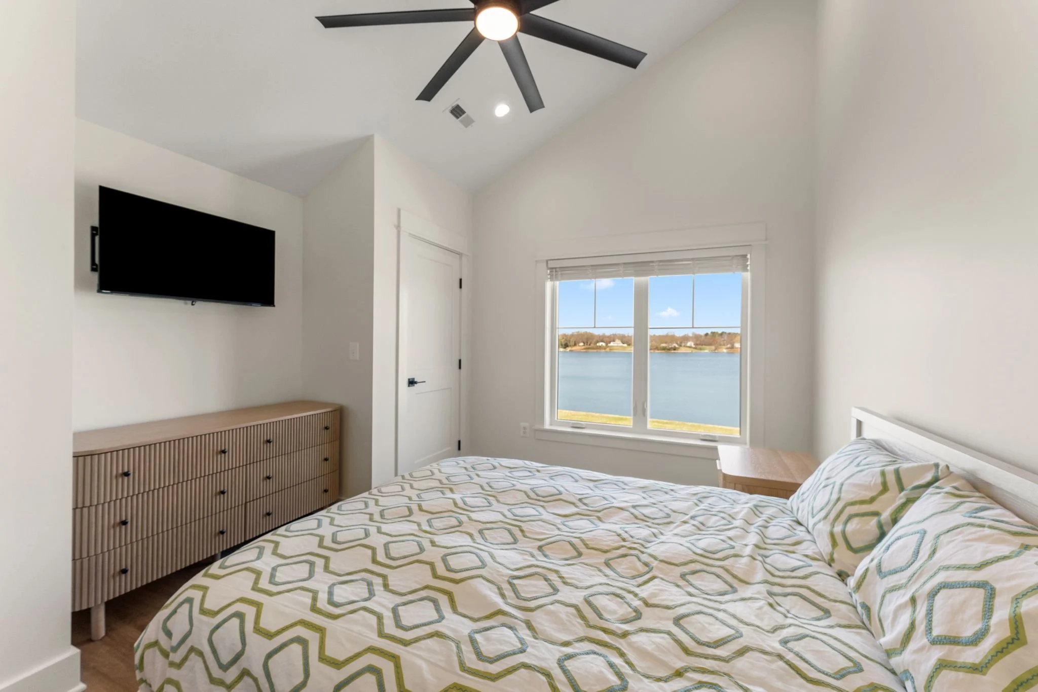 Upstairs bedroom with a large window overlooking the water, a queen-sized bed, a wooden dresser, a wall-mounted TV, and a ceiling fan.