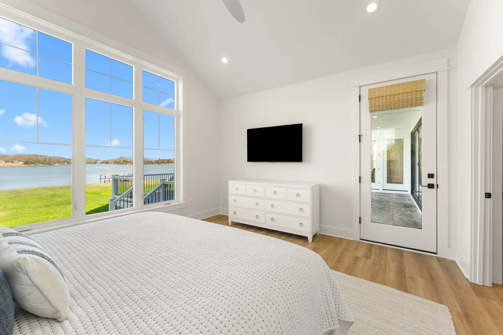 Bright master bedroom with large windows overlooking the water, king-sized bed, a white dresser, a wall-mounted TV, and a glass door leading to the retractable screened porch.