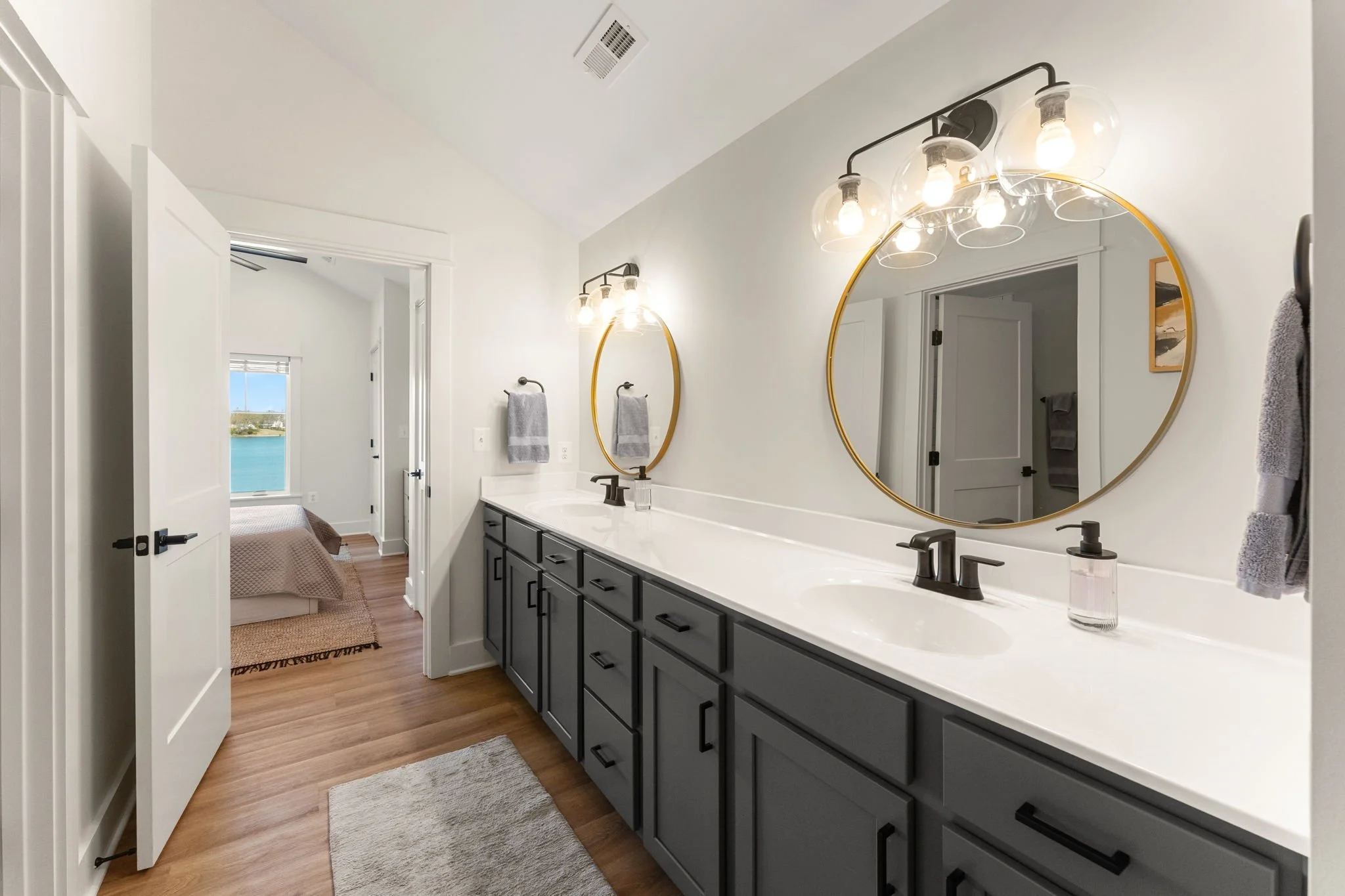 Upstairs bathroom with dual vanities, large round mirrors with gold frames, and modern light fixtures. View into a bedroom with a window overlooking water.