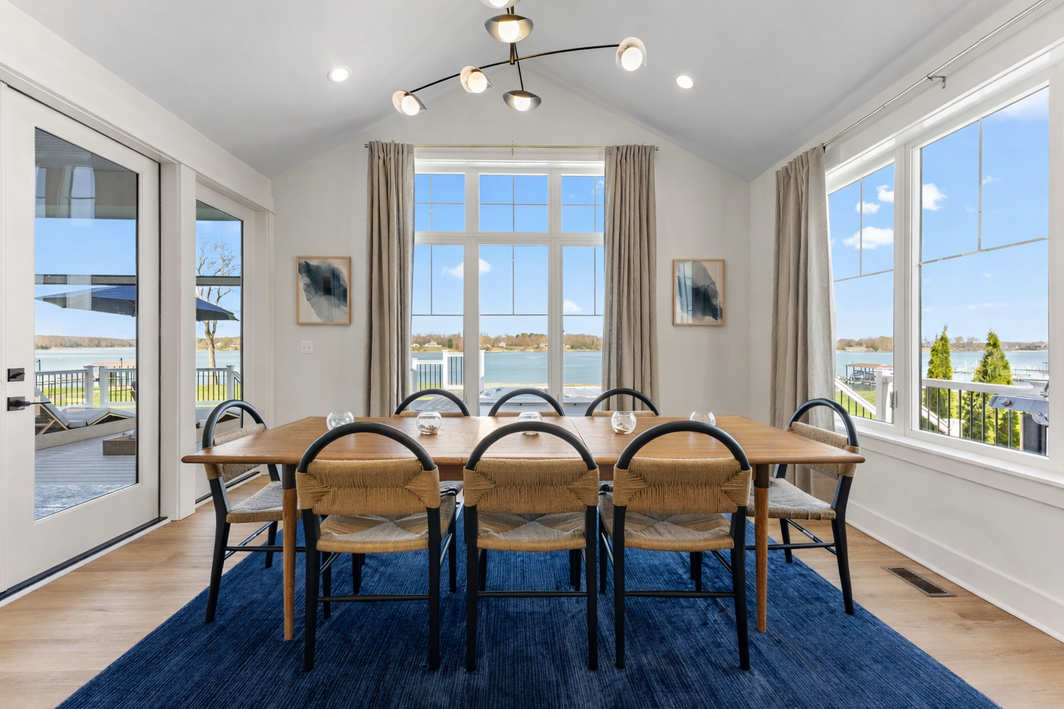 A dining room with a large wooden table, eight chairs, and a view of the water through large windows.