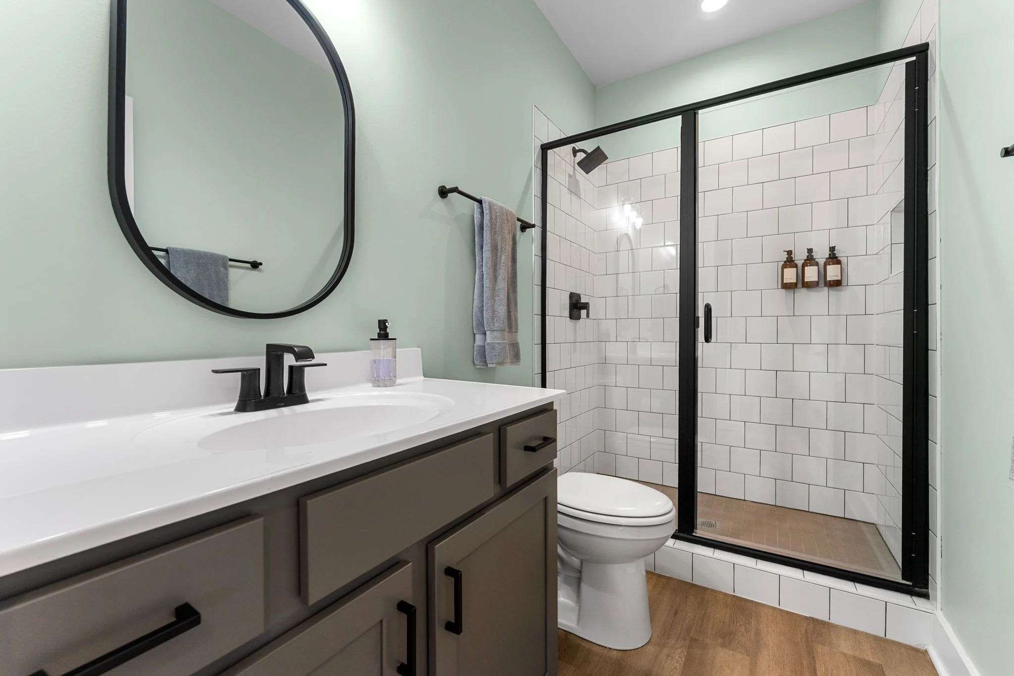 Bathroom with sea salt green walls, black-framed mirror, glass shower with black frame, and shampoo/conditioner/soap.