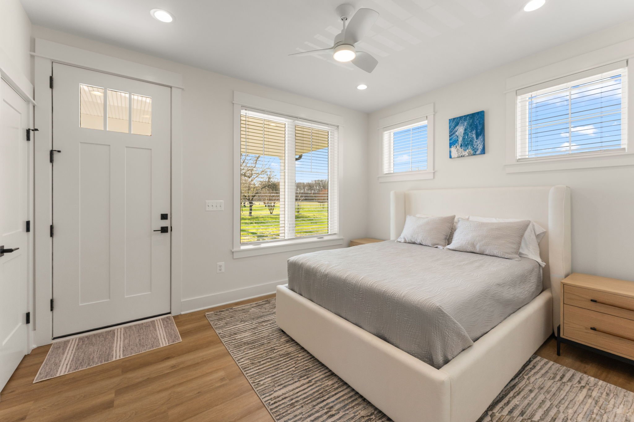 Bright bedroom with white walls, a queen bed, wooden nightstands, and windows facing the front yard.