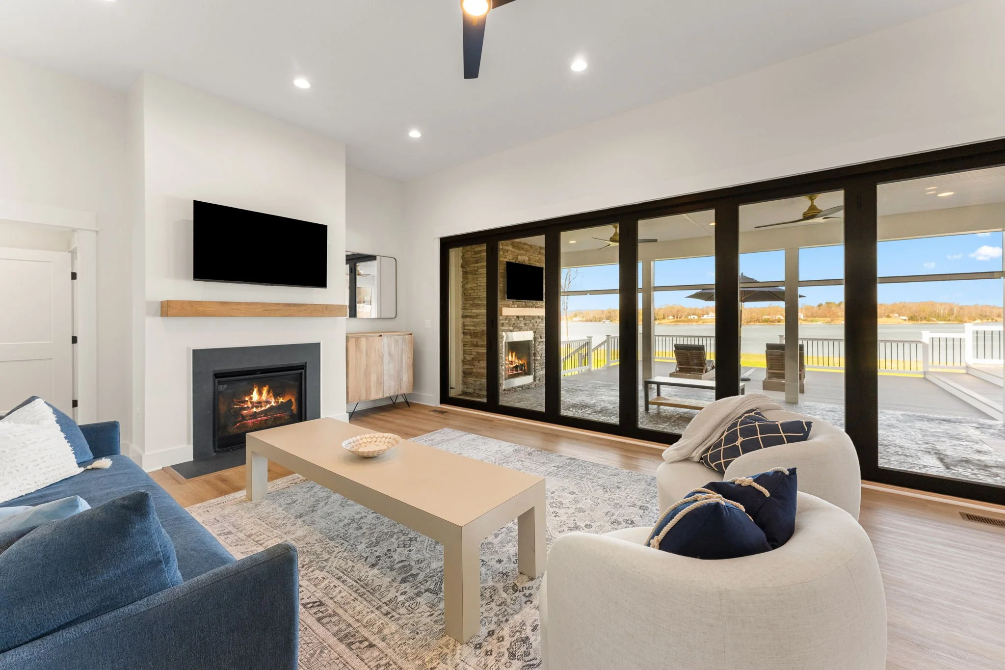Living room with a fireplace, a television mounted above, and a fully collapsible sliding glass wall leading to an outdoor patio with retractable screens and a waterfront view.