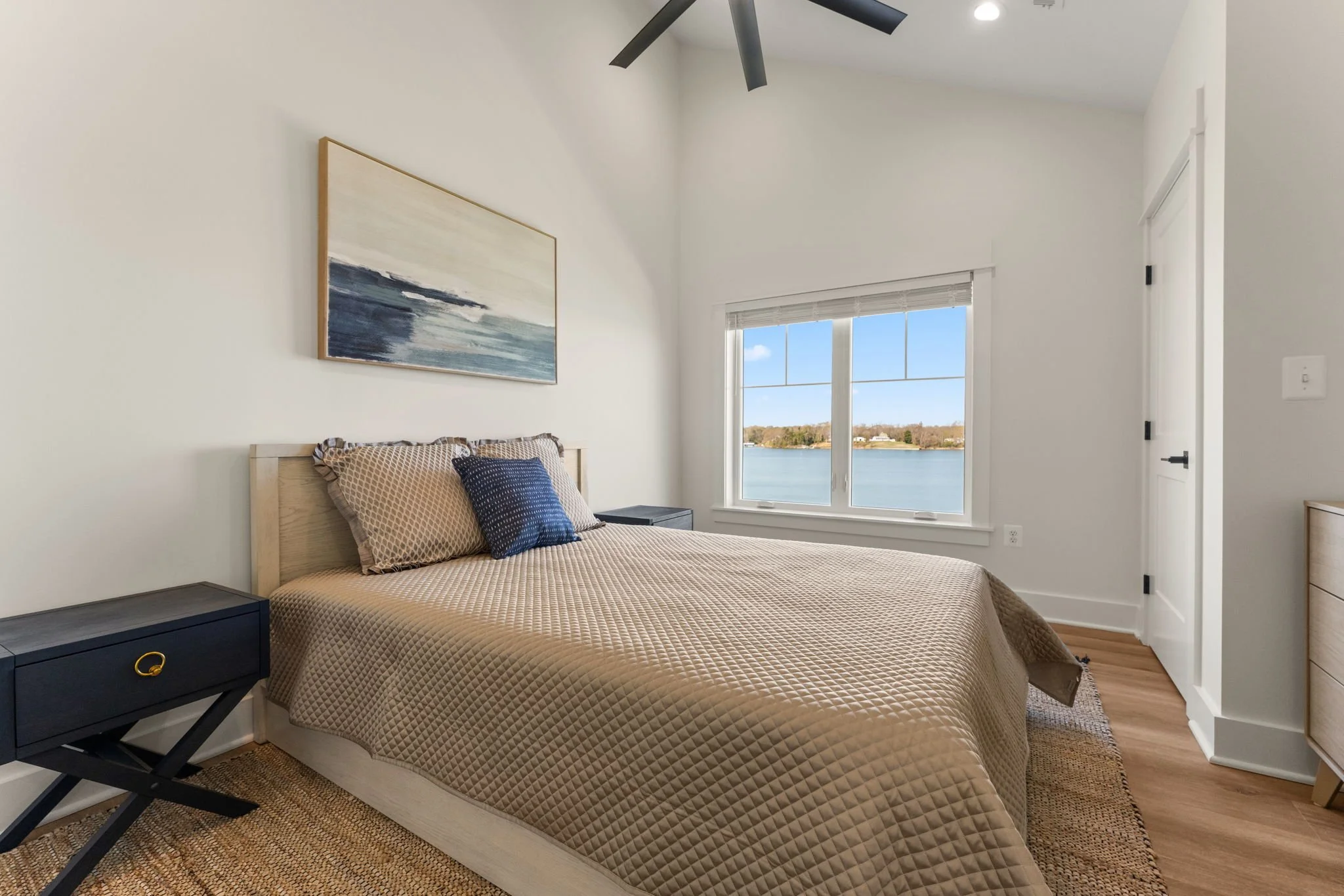 Upstairs bedroom with bedside tables, large windows facing the water, white walls, and a ceiling fan.