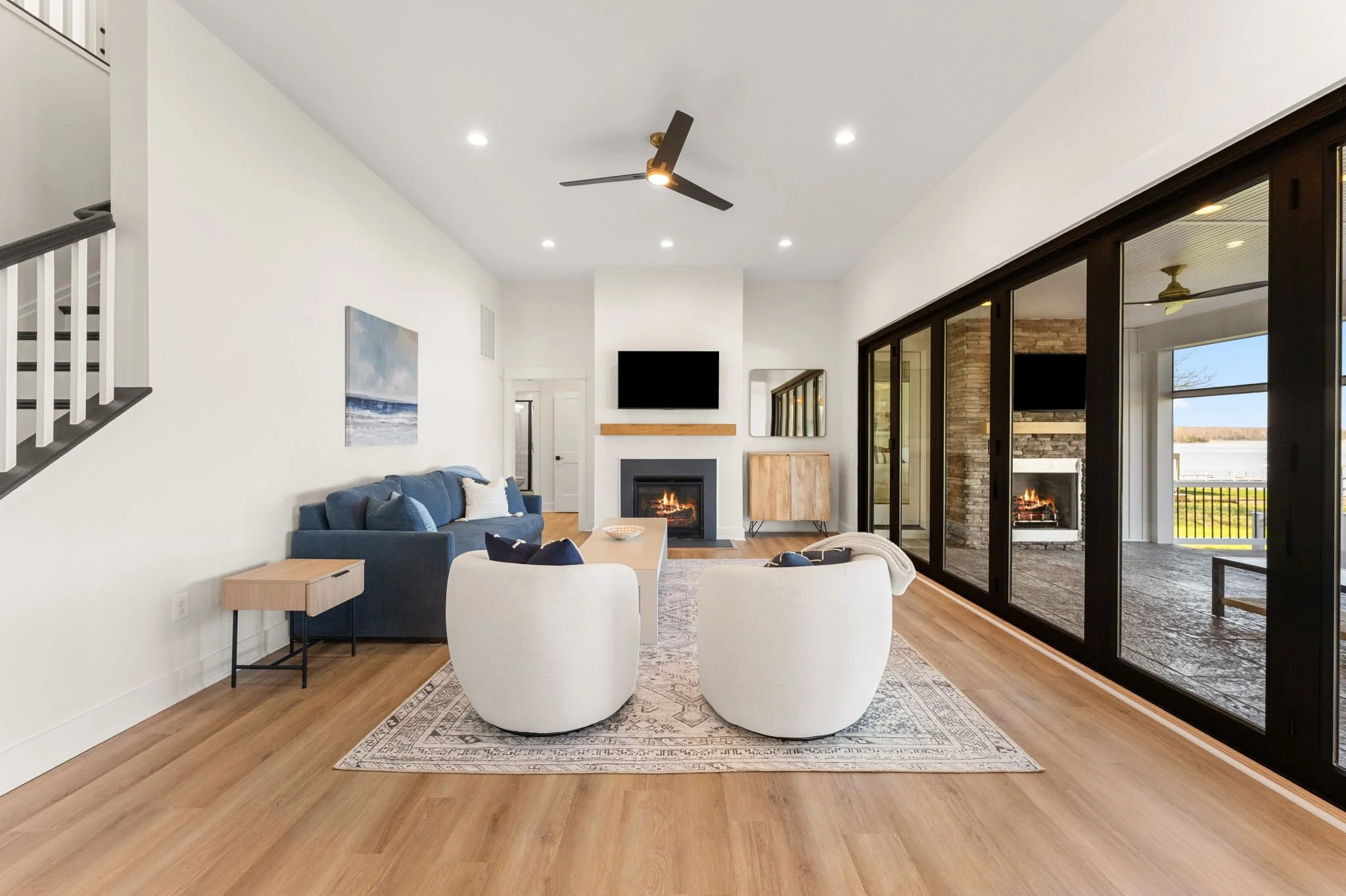 Modern living room with sofa, chairs, a fireplace, a wall-mounted TV, and a fully collapsible sliding glass wall leading to an outdoor patio with retractable screens and a waterfront view.