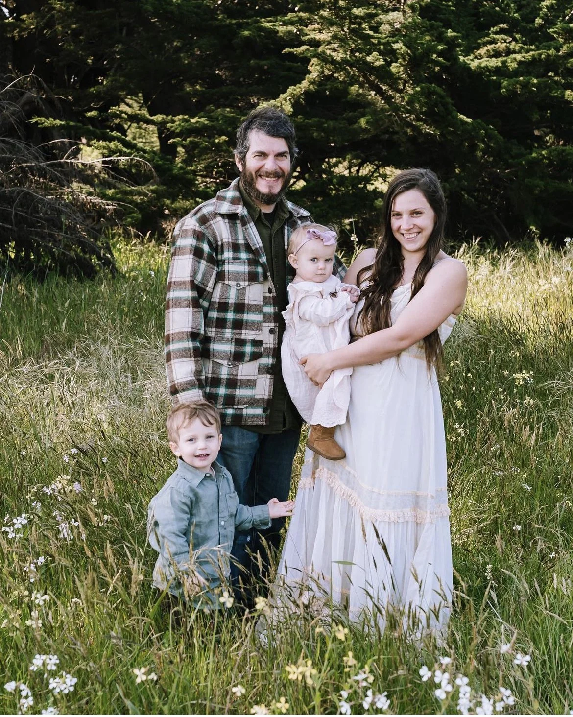 A family of four standing in a grassy field with trees in the background. The father has dark hair and a beard, wearing a plaid jacket. The mother has long dark hair, wearing a white dress. The daughter is being held by the mother, wearing a pink dress and bow. The son, wearing a light blue shirt, stands in front of them. All are smiling and looking at the camera.