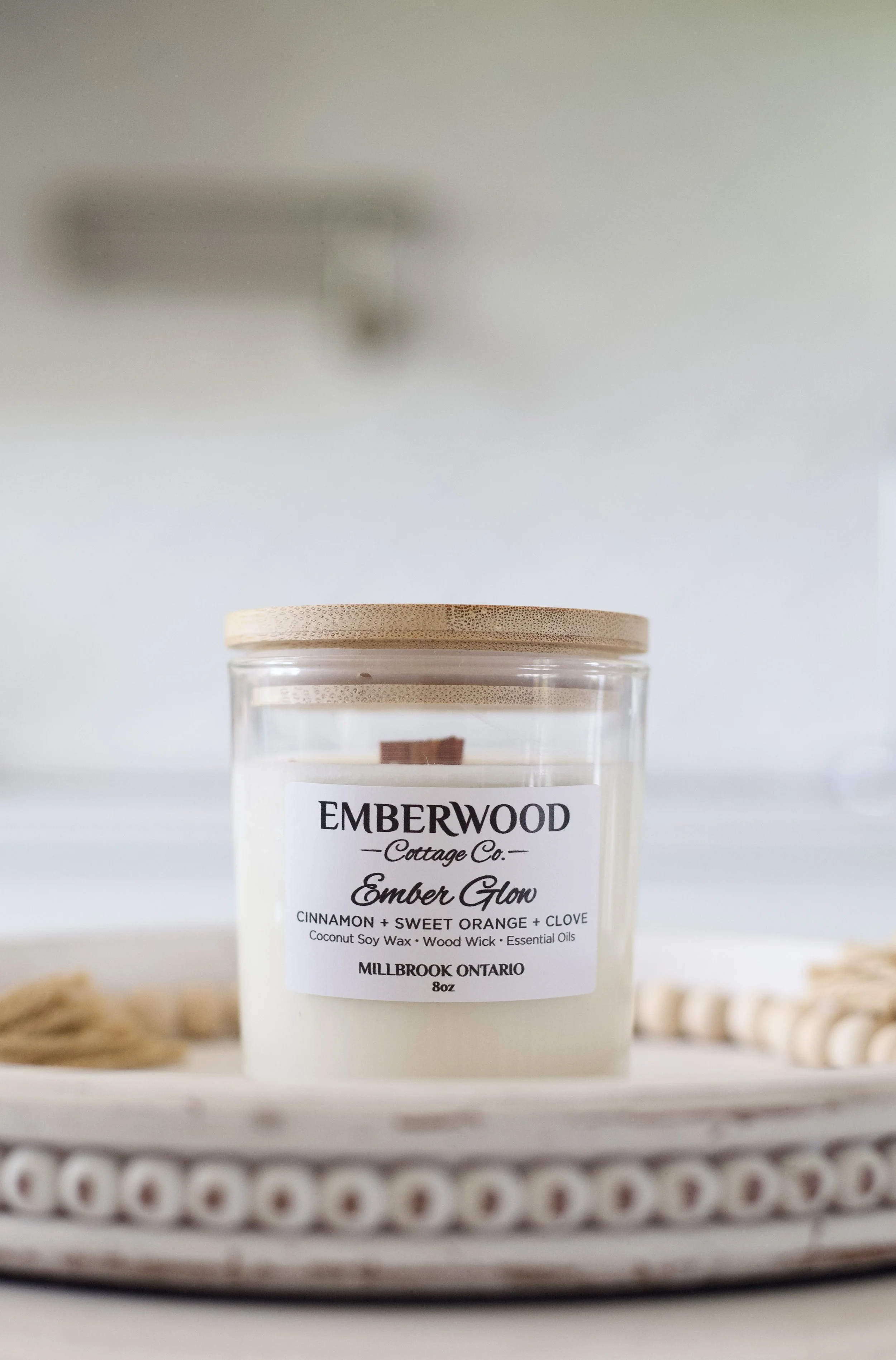 A candle labeled 'EmberGlow' from EmberWood Cottage Co. with scents of cinnamon, sweet orange, and clove in a jar with a wooden lid, placed on a white surface with a blurred background.