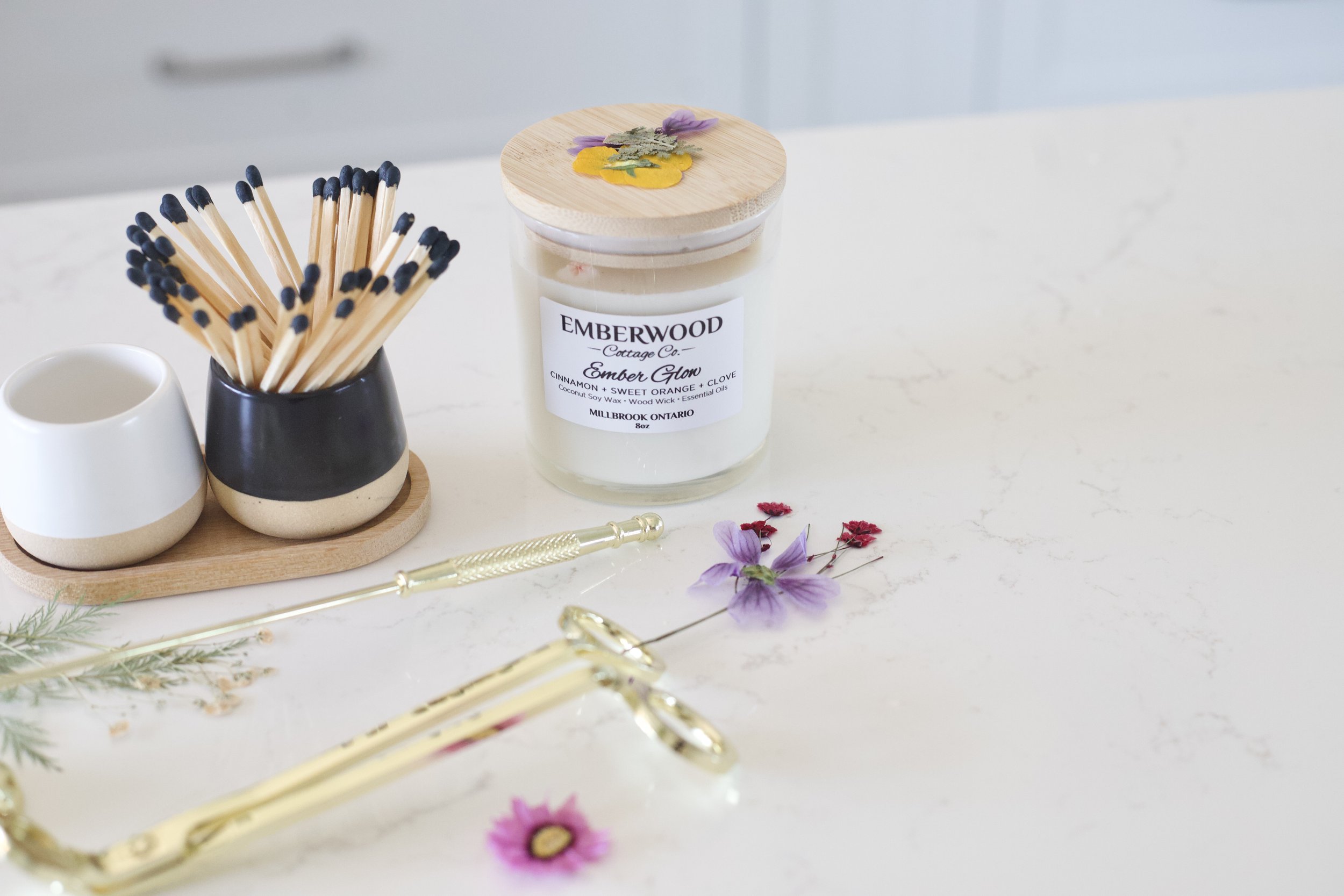 A white marble surface with decorative items including a black and beige ceramic cup holding matches with black tips, a small beige and white ceramic bowl, a wooden tray, a candle labeled 'Ember Glow' with a wooden lid and dried flower decoration, some gold-colored jewelry, and small purple and pink dried flowers.