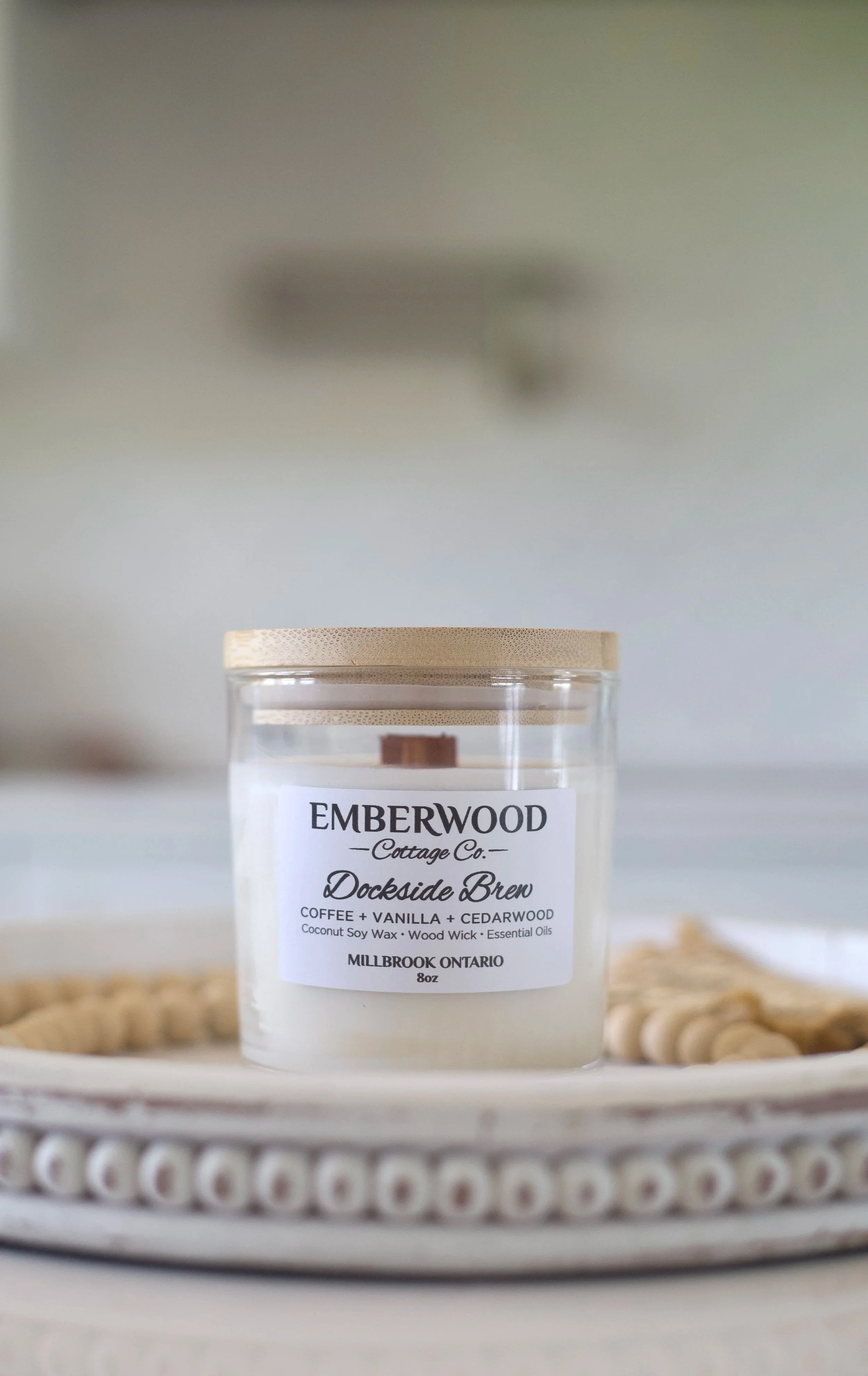 A candle labeled 'Emberwood Cottage Co. Docksde Bren' with scents of coffee, vanilla, and cedarwood, in a small glass jar with a wooden lid, placed on a tray with decorative beads.
