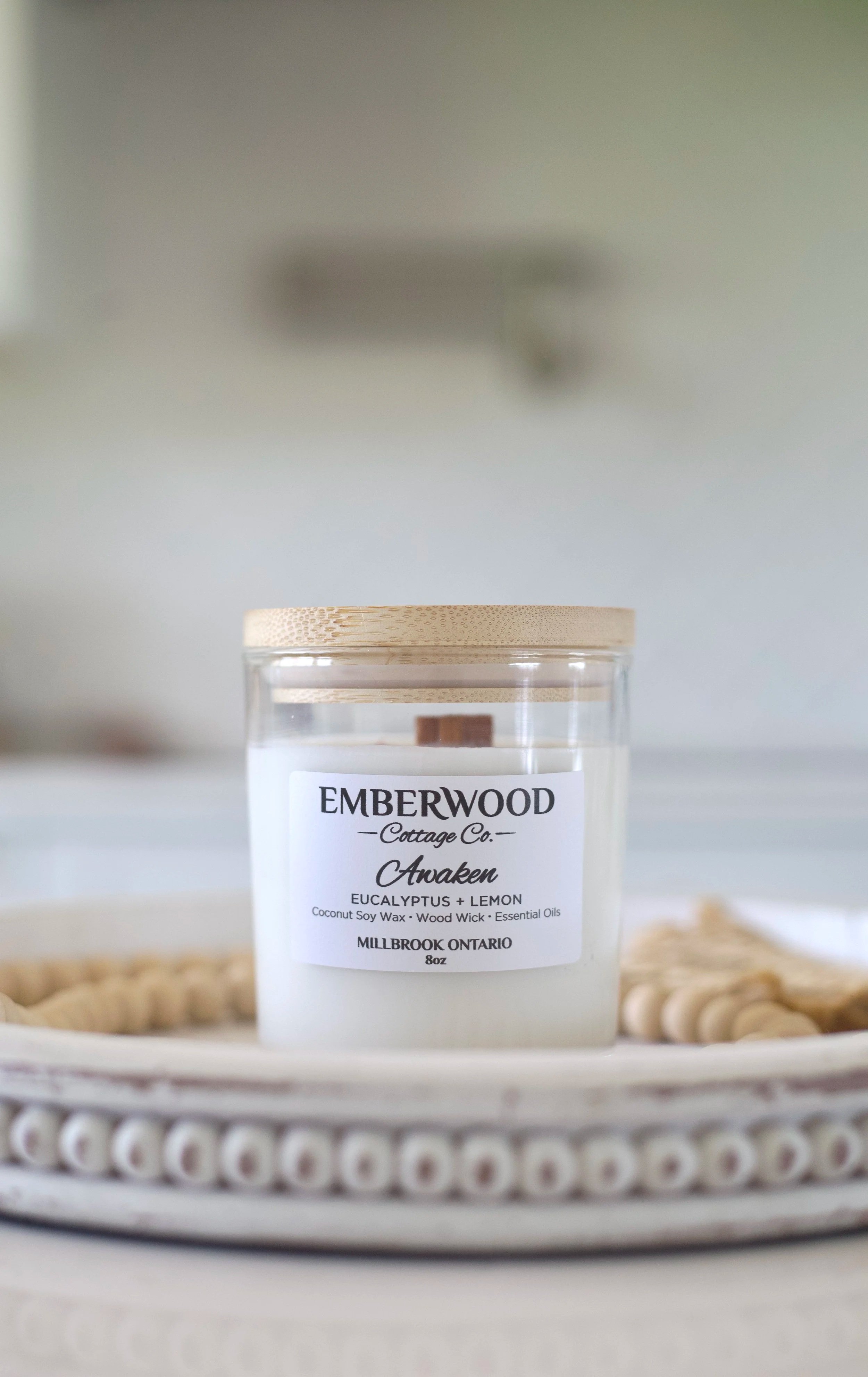 A white candle with a wooden lid labeled 'EMBERWOOD Cottage Co. Awaken, Eucalyptus + Lemon, Coconut Soy Wax, Wood Wick, Essential Oils, Millbrook Ontario, 8oz' on a decorative tray with cream-colored beads.