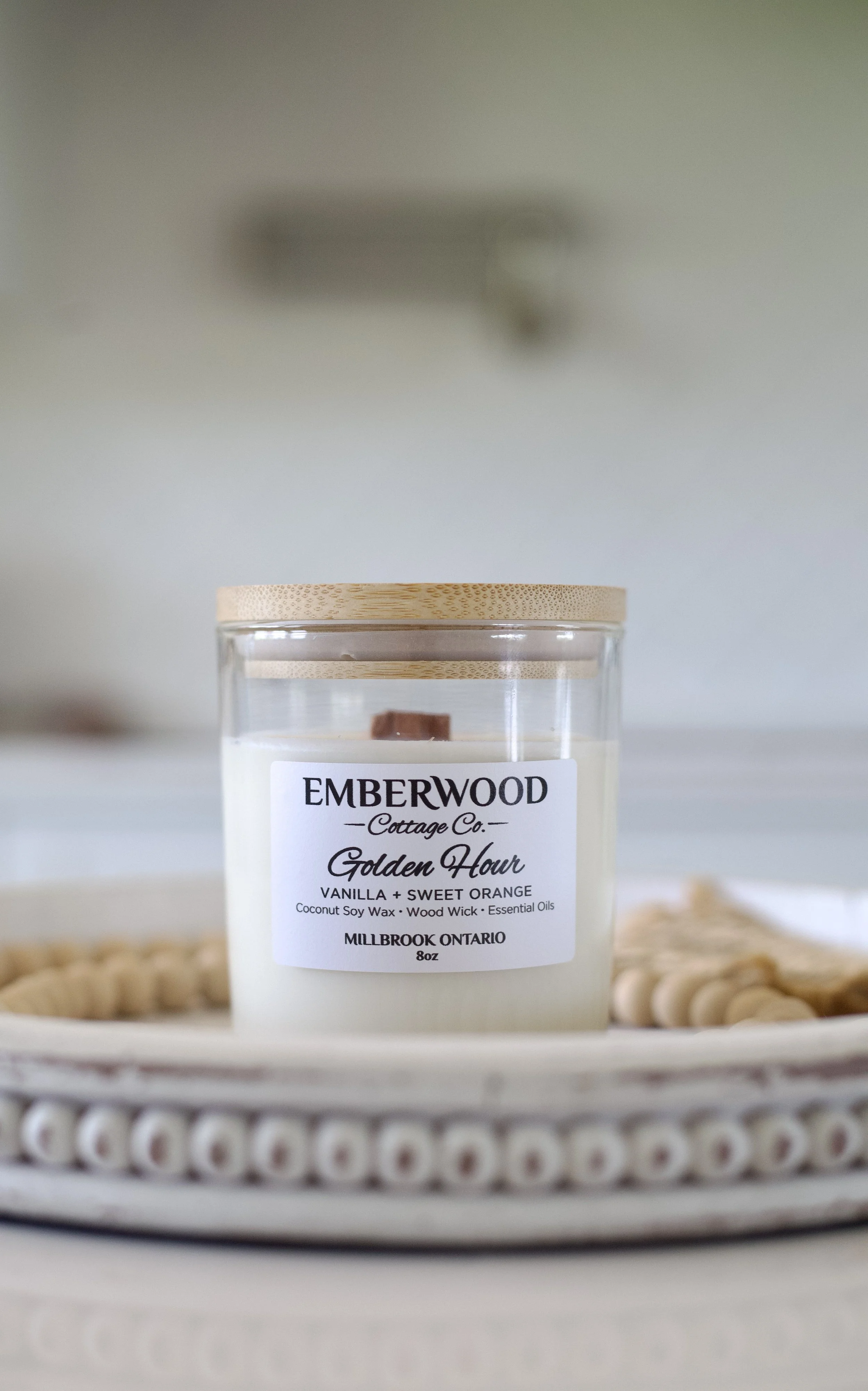 Close-up of a white candle with a wooden lid and a label that reads 'Emberwood Cottage Co. Golden Hour Vanilla + Sweet Orange,' placed on a decorative tray with a beaded edge.