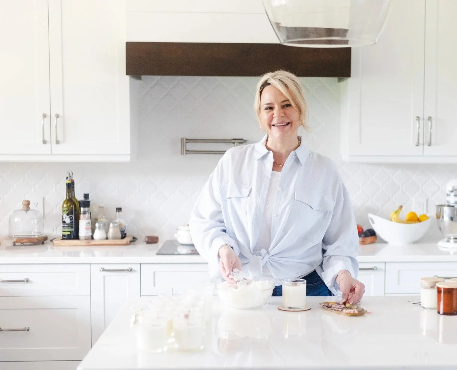 Krista the owner smiling in a white kitchen, wearing a white shirt with a knot at the waist. She is standing behind a counter with bowls and glasses.