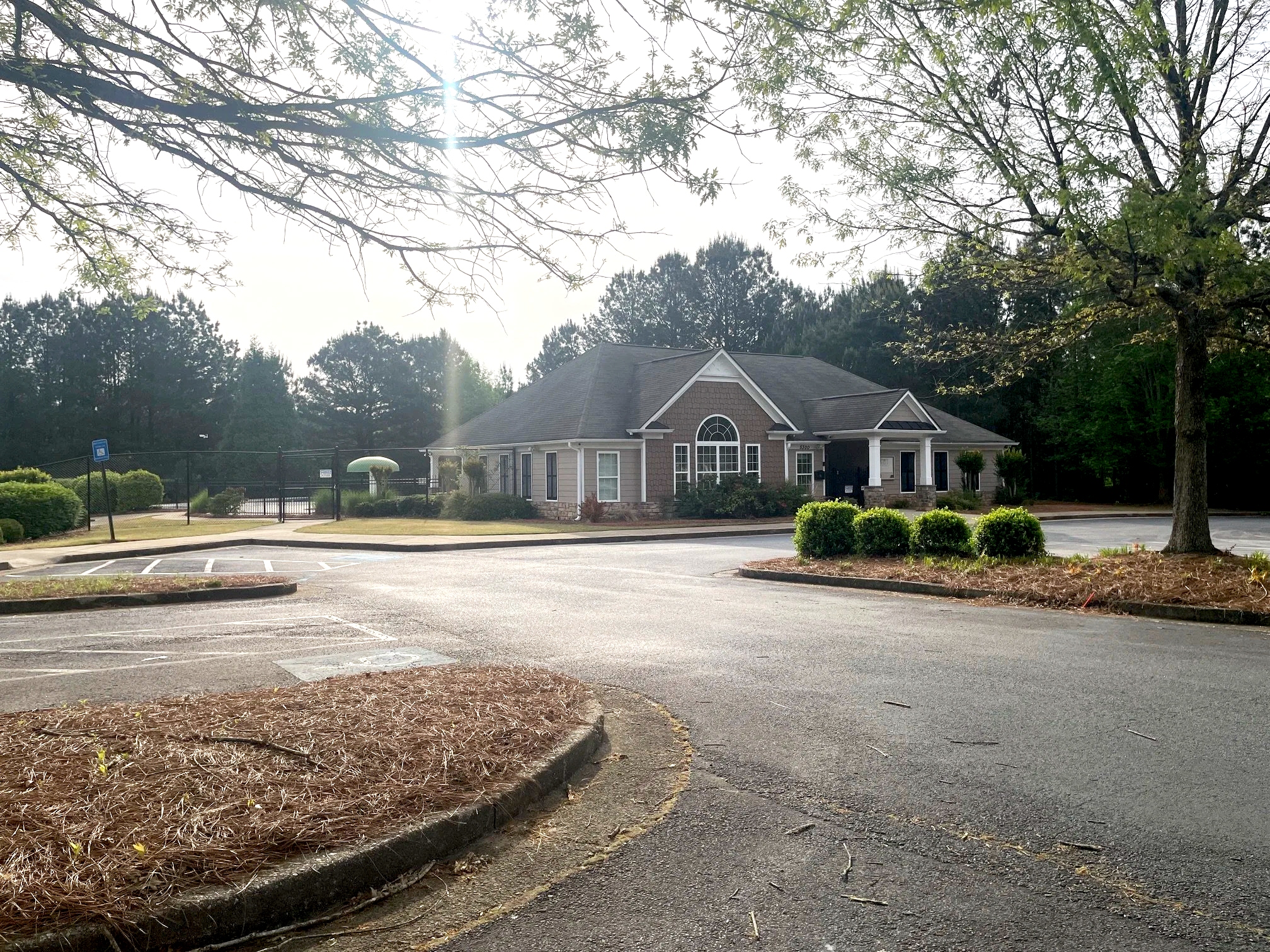 A residential area with a house, parking lot, trees, and shrubs under a bright sky with sunlight filtering through tree branches.
