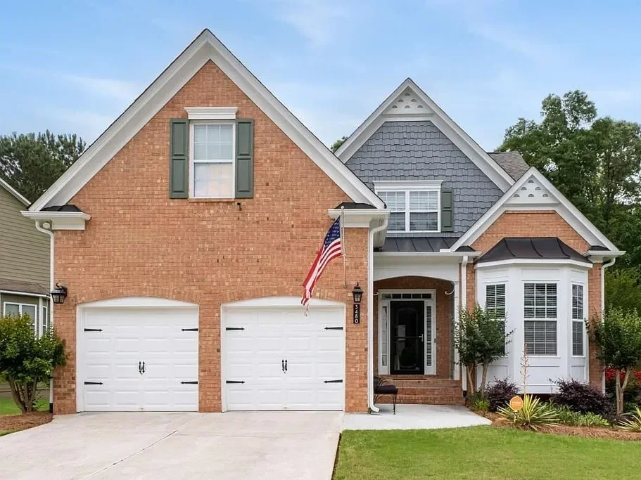Front view of a two-story house with a brick facade, white garage doors, and an American flag near the entrance. The house has a combination of brick and gray siding, with green shutters and a small front porch with steps.