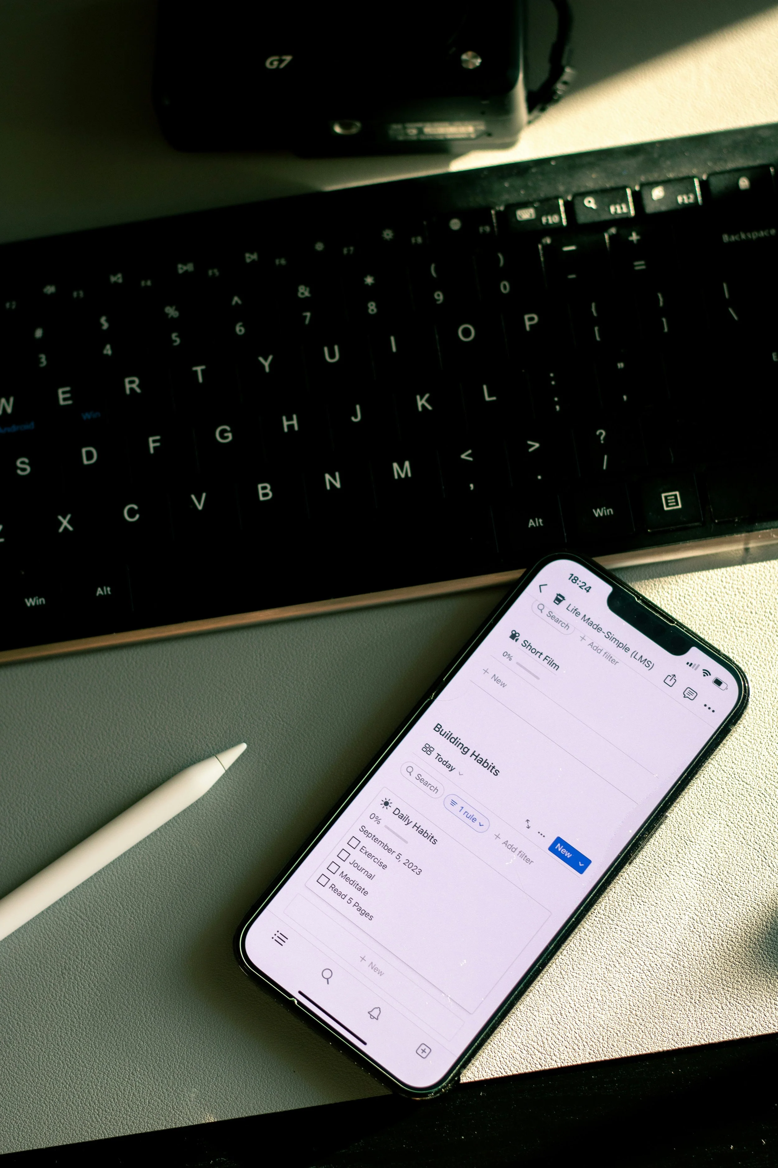 A smartphone screen displaying a to-do list app with items such as 'Building Habits' and 'Daily Habits', a white pen, a computer keyboard, and a camera in the background on a desk.
