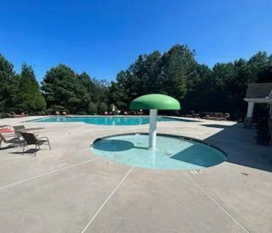 Empty swimming pool with a large green umbrella-shaped water feature and patio lounge chairs, surrounded by trees under a clear blue sky.