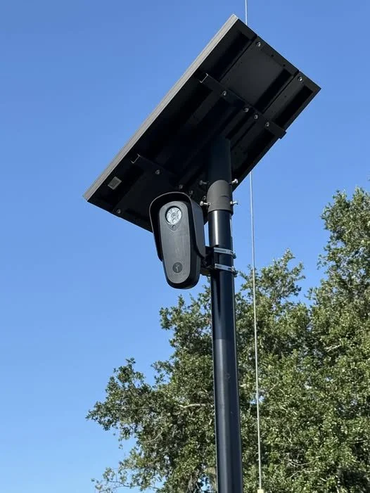 Solar-powered surveillance camera mounted on a pole against a clear blue sky with trees.