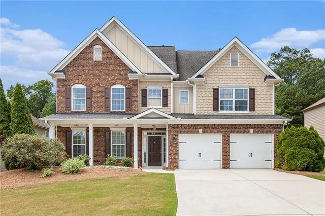 A two-story suburban house with a brick and beige siding exterior, brown shutters, a covered front porch, three garage doors, a manicured lawn, and some trees and shrubs surrounding it.