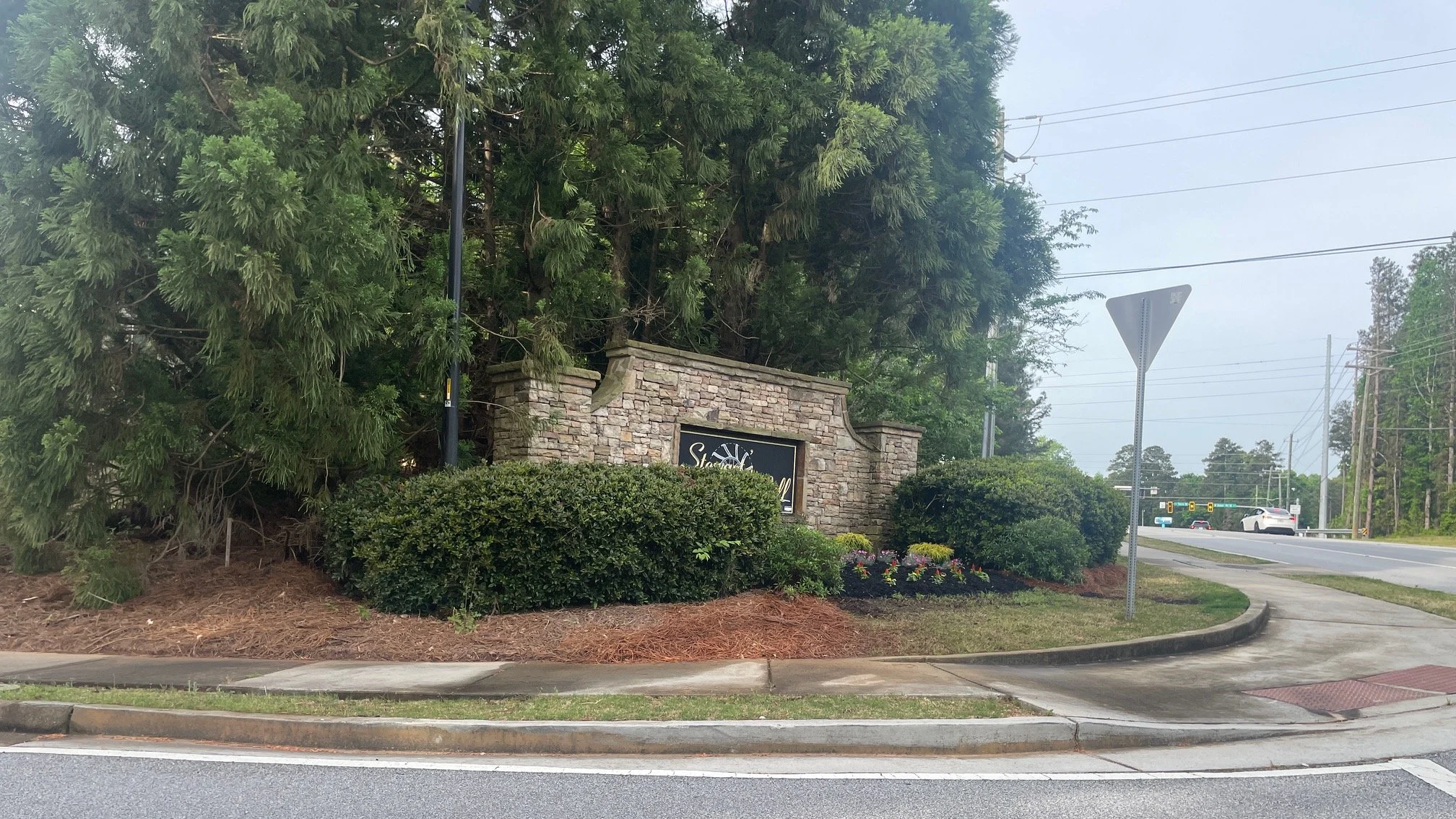 A stone sign with the text 'Stone Valley' surrounded by bushes and trees, near a road with cars and traffic lights in the background.