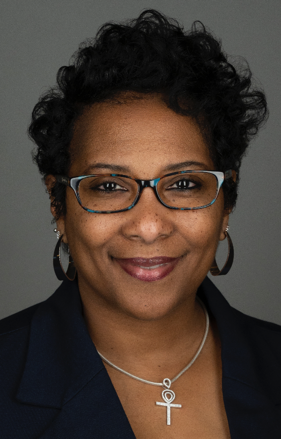 A woman with short curly hair, glasses, and hoop earrings, smiling in front of a gray background.