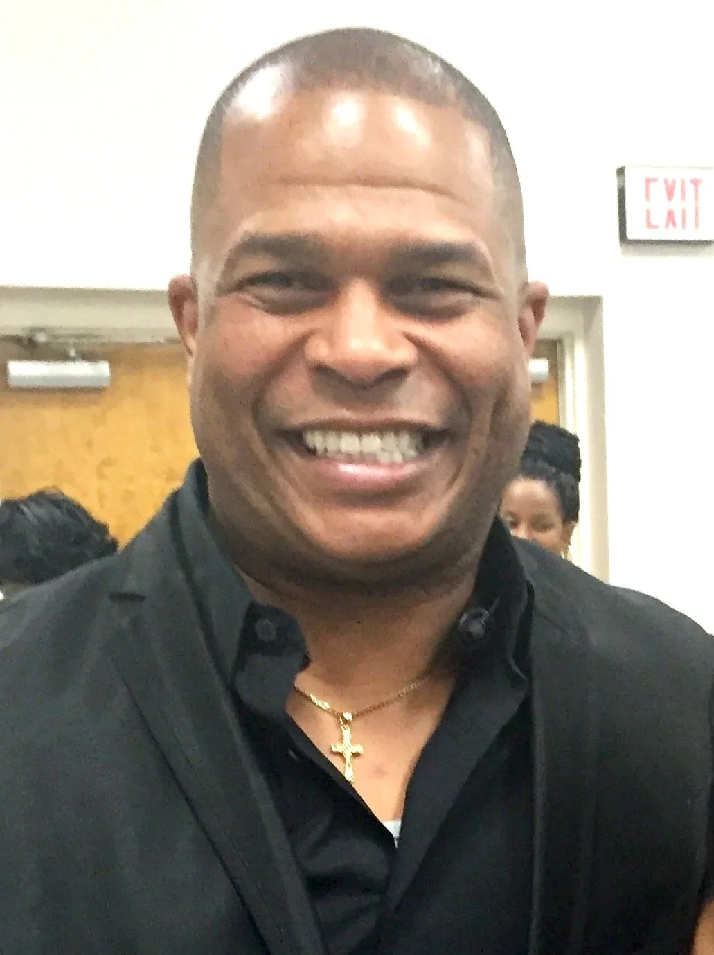A close-up of a smiling man with a gold cross necklace, wearing a black shirt, at an indoor event.