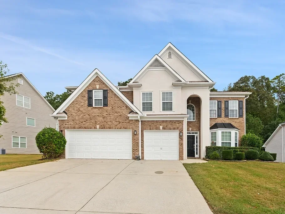 A large, two-story house with a combination of brick and white siding, three garage doors, and multiple windows. The house has a front yard with grass and bushes and is set under a blue sky.