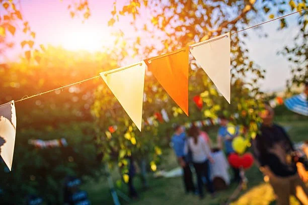 String of triangular bunting flags hanging outdoors at sunset during a celebration, with blurred people and decorations in the background.