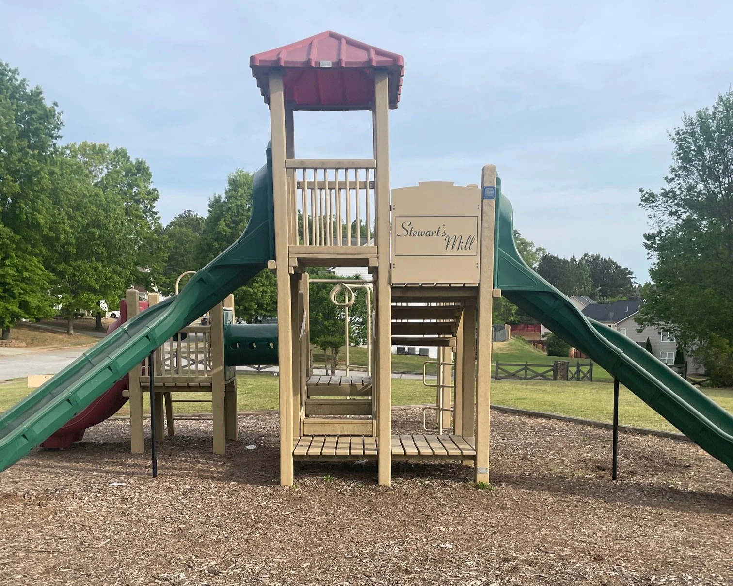 Playground structure with slides, ladders, and a small platform, set in a park with grass, trees, and houses in the background.