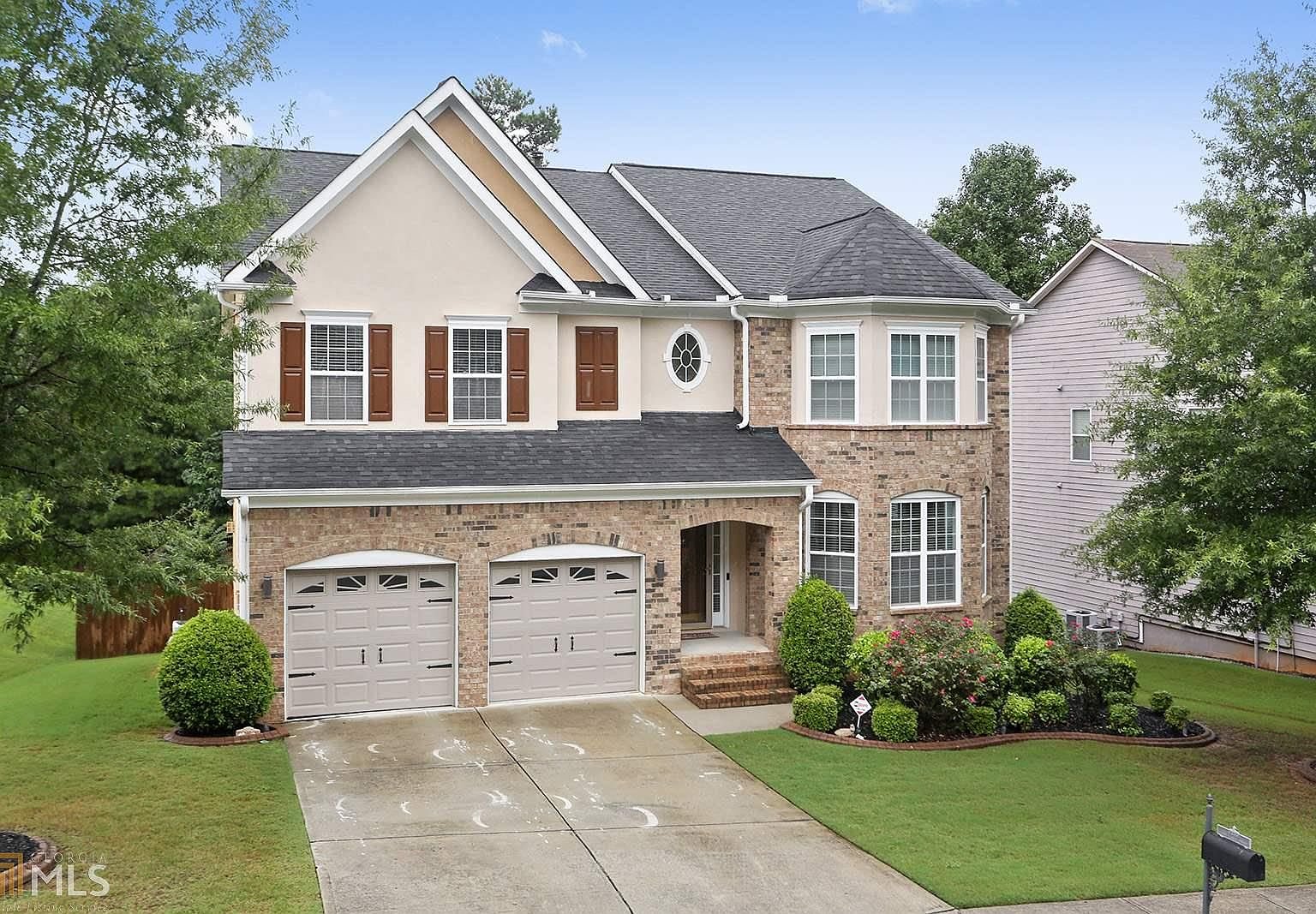 A two-story suburban house with brick and beige siding, two white garage doors, a small front porch, and landscaped garden with bushes and flowers, under a blue sky.
