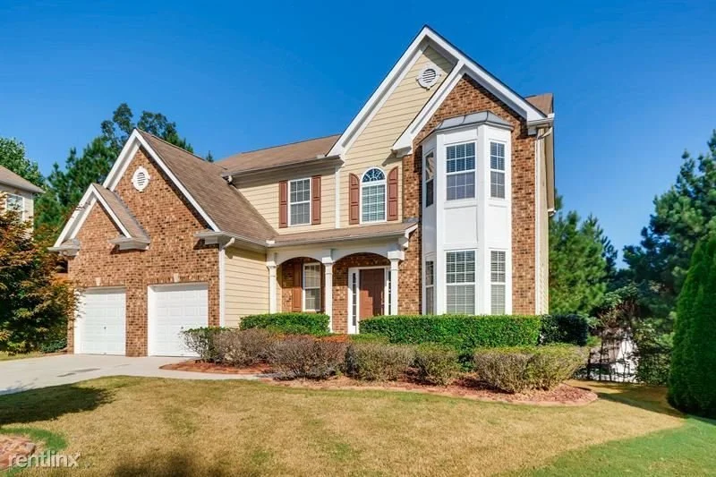 Two-story house with brick and beige siding, large bay windows, brown shutters, and a two-car garage, surrounded by a well-maintained lawn and bushes, under a bright blue sky.