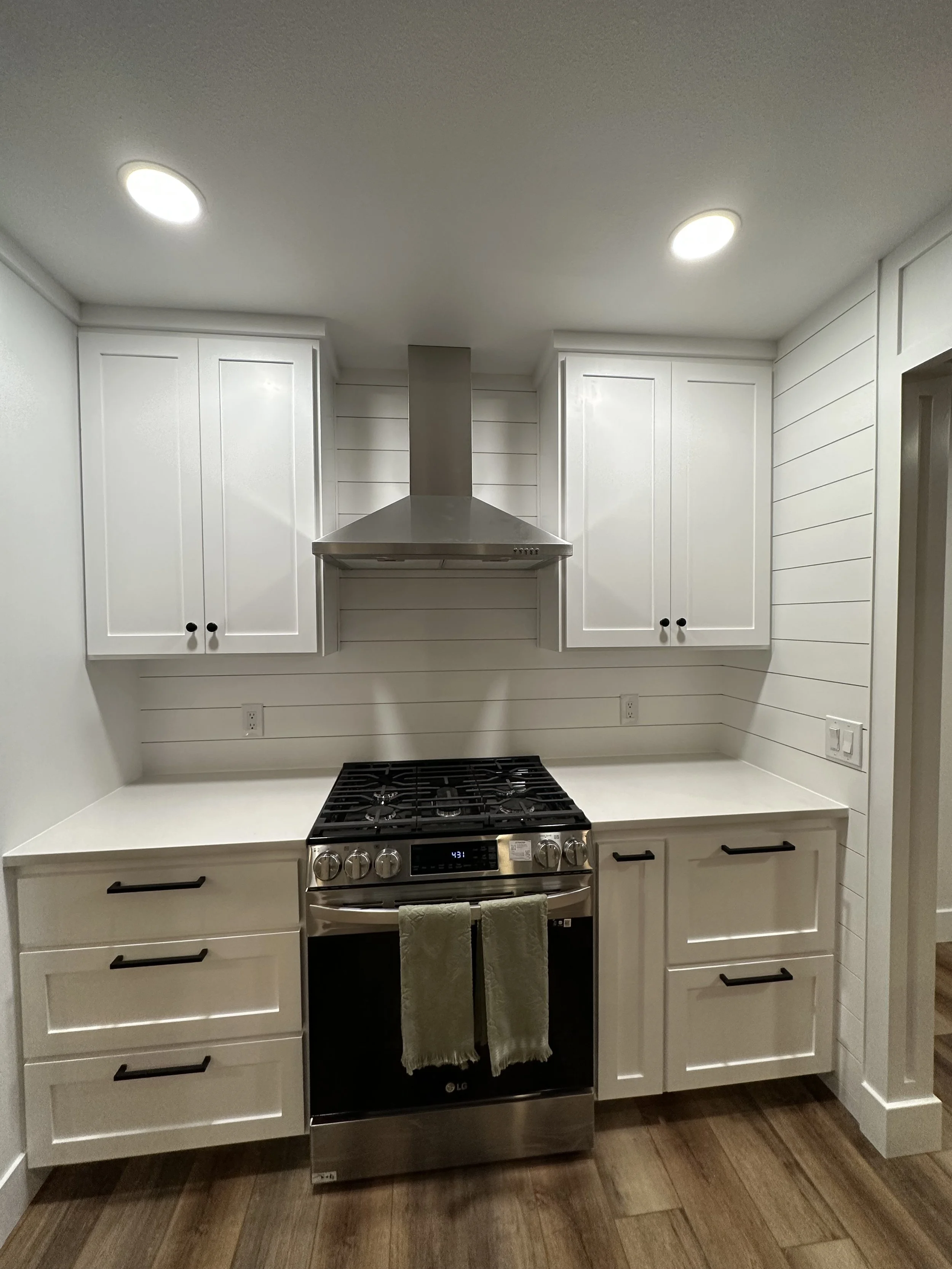 Modern kitchen with white cabinets, stainless steel oven and range hood, wood flooring, and recessed ceiling lights.